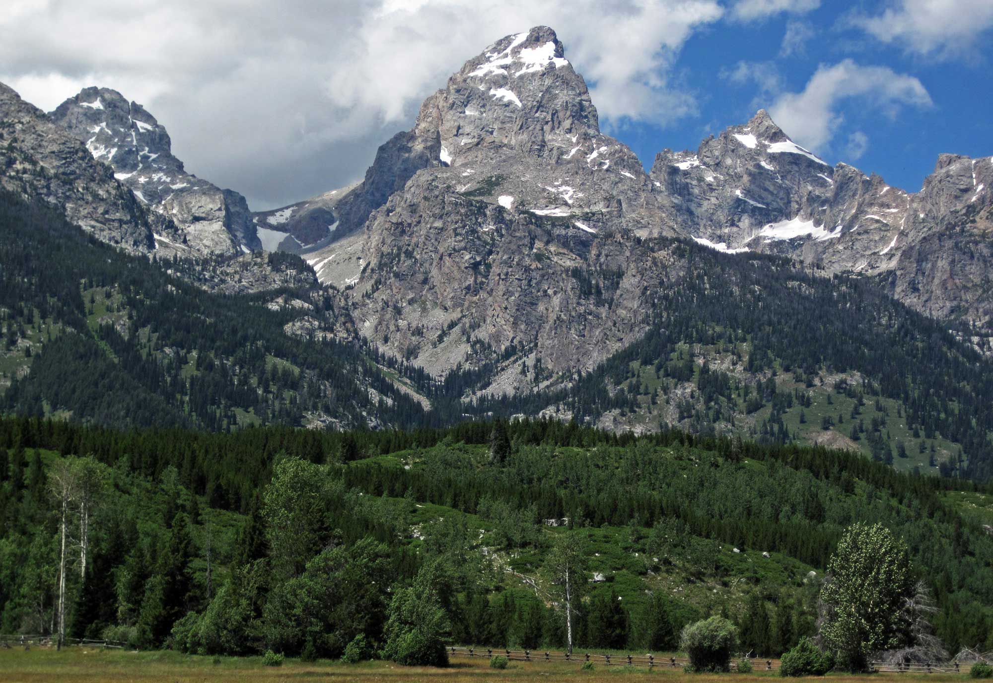 Photograph of Grand Tetons, Wyoming. The photo shows a rugged, triangular mountain peak made of gray rock in its center with additional shorter peaks flanking it. The rocky peaks have patches of snow. The slopes at lower elevation are covered with conifers. A but of a flat field can be seen in at the bottom foreground of the image.