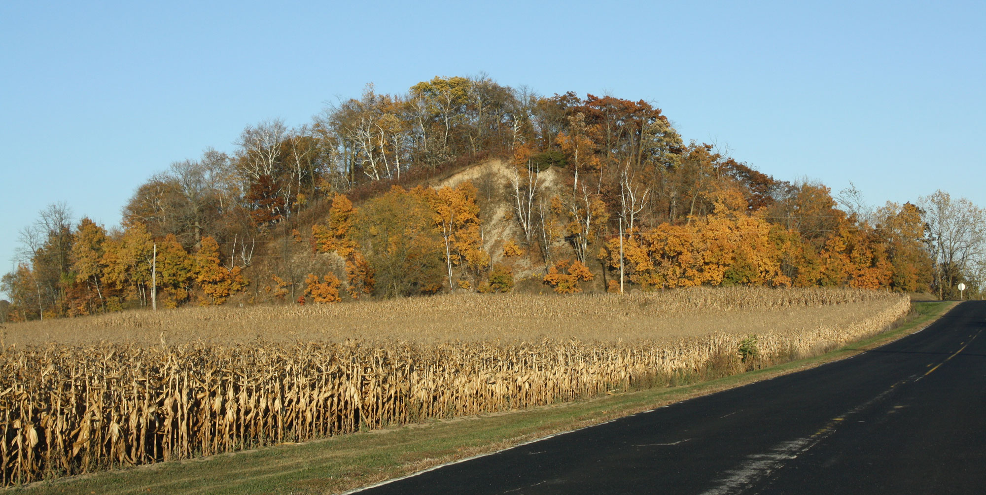 Photograph of a kame in Wisconsin. The photo shows a field of dried cornstalks and a paved road in the foreground. A rounded hill covered with trees rises in the background. 