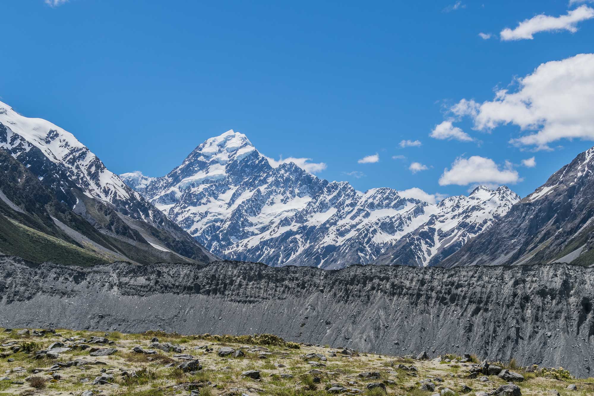 Photograph of Mueller lateral moraine in New Zealand. The photo shows a gray ridge running horizontally across the foreground of the image, which is the moraine. A snow-dusted mountain rises in the background.