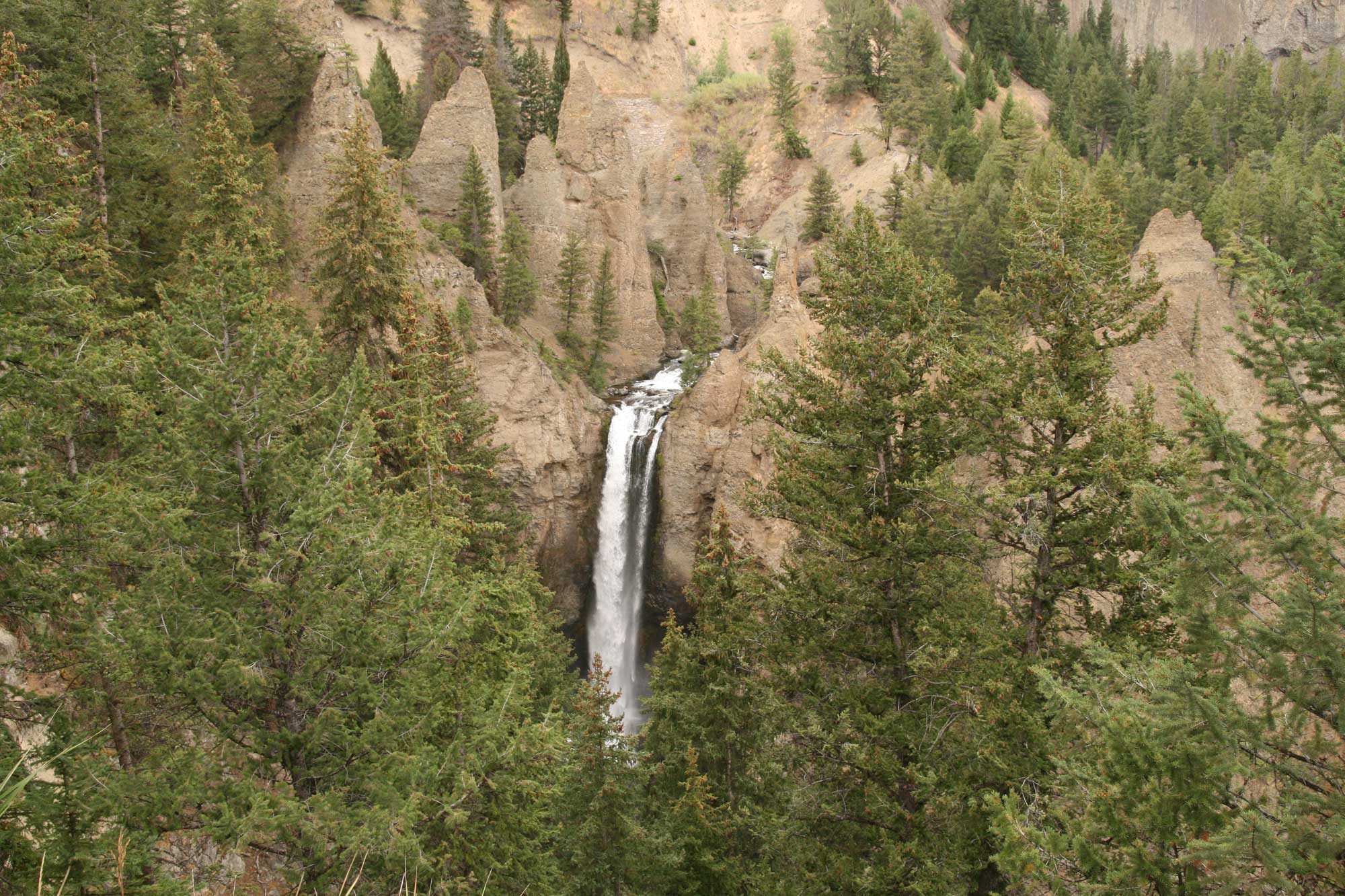 Photograph of Tower Fall, Yellowstone National Park. The photograph shows a tall, narrow waterfall dropping from a cliff at the center of the image. Brown rocky peaks rise around it. The landscape is dotted with conifers.