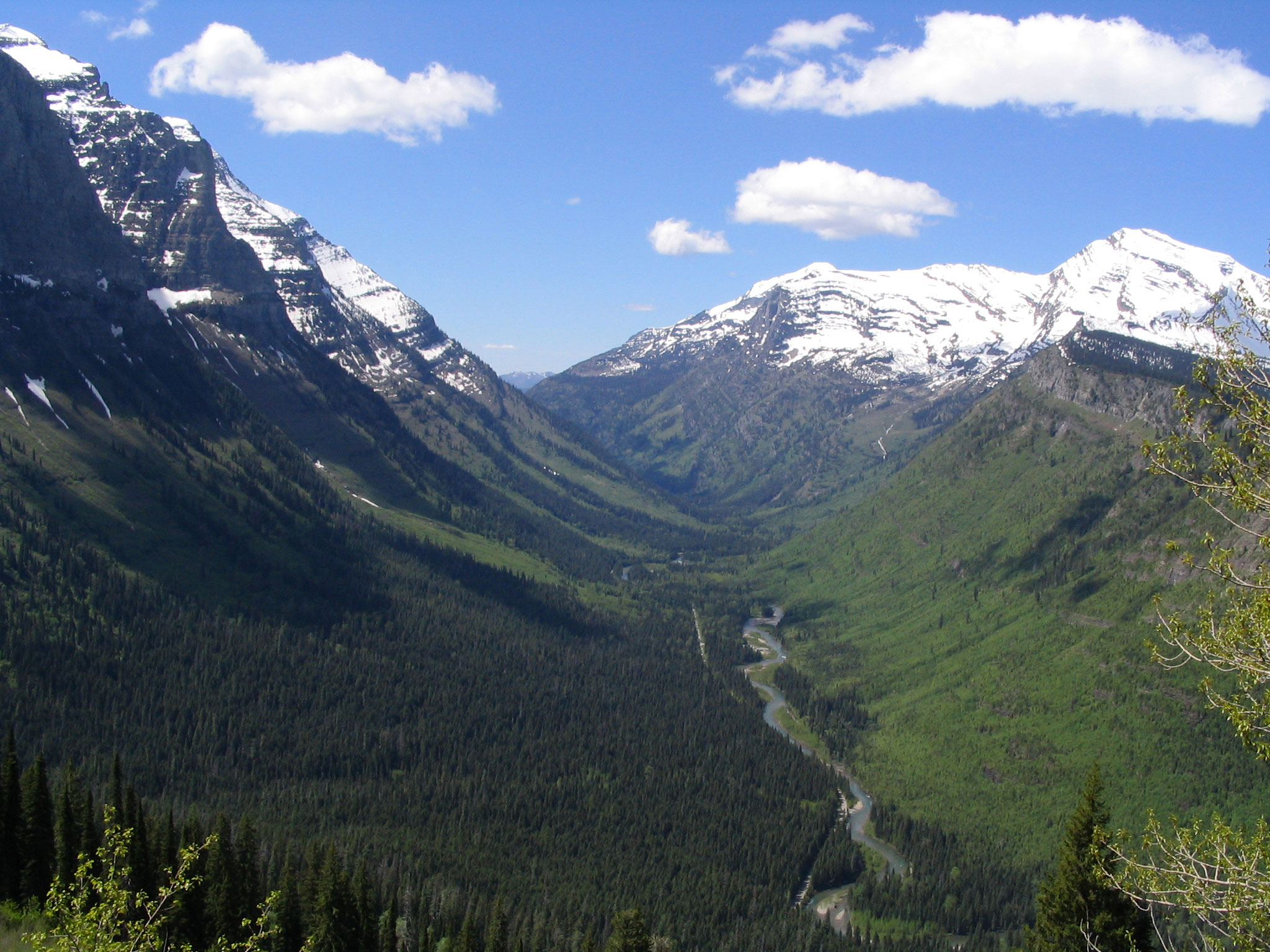 Photograph of a U-shaped, glacially carved valley in Glacier National Park, Montana. The photo shows a broad, u-shaped valley with snow-dusted mountain slopes rising on the left and snow-dusted mountain peaks in the background at right. At lower elevations, the slopes are vegetated.
