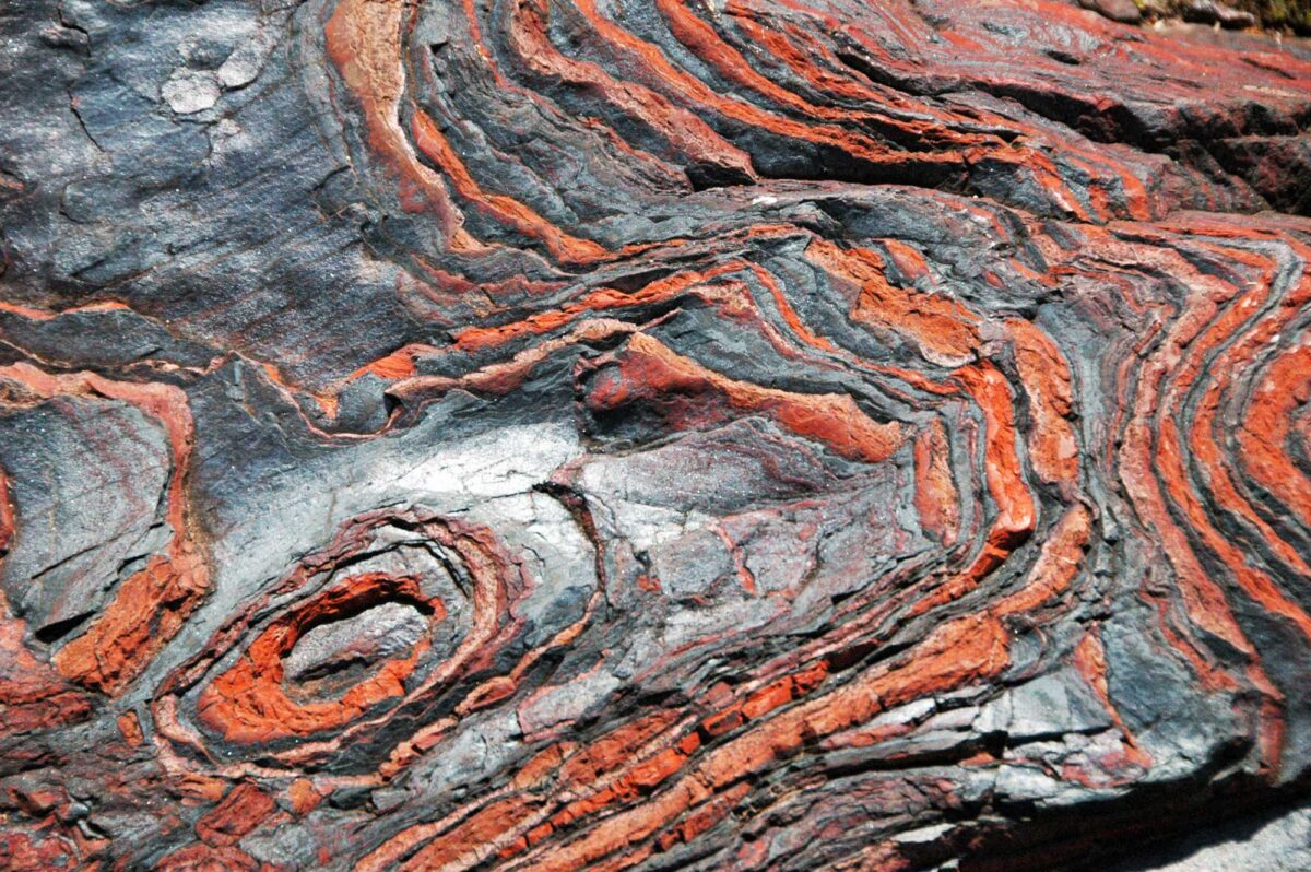 Photograph of banded iron from the Paleoproterozoic of Michigan. The photo shows the surface of a rock that is dark gray with red-orange layers in it. The layers are sinuous.