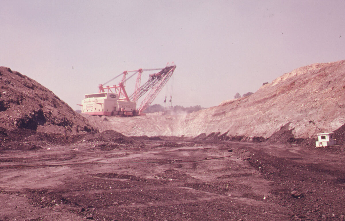 Photograph of Big Muskie, a large dragline excavating coal in a strip mine in Ohio in 1974. The photo shows a red and while machine with arms at the front, from which a large bucket is suspended by cables. A partially excavated hill is in front of the dragline. Behind it is a pile of earth or coal.