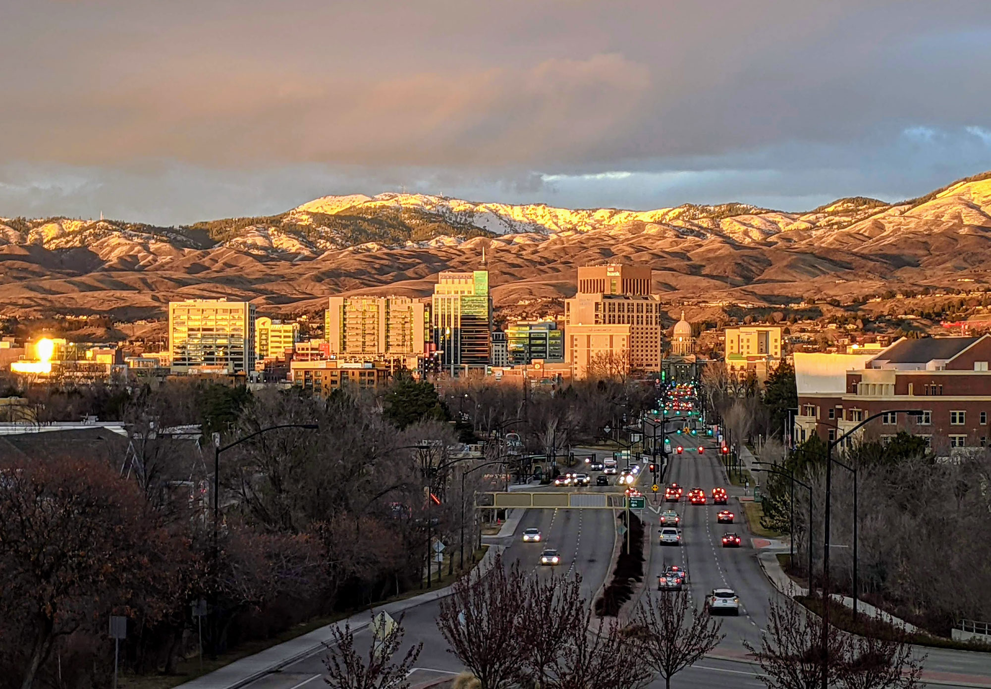 Photograph of Boise, Idaho, in 2021, possibly at sunset. The photo shows a city with short skyscrapers clustered in the mid-distance. Paved three-lane roads running to and from the city center can be seen in the foreground. In the background, snow-dusted rolling hills can be see. The sky is overcast.