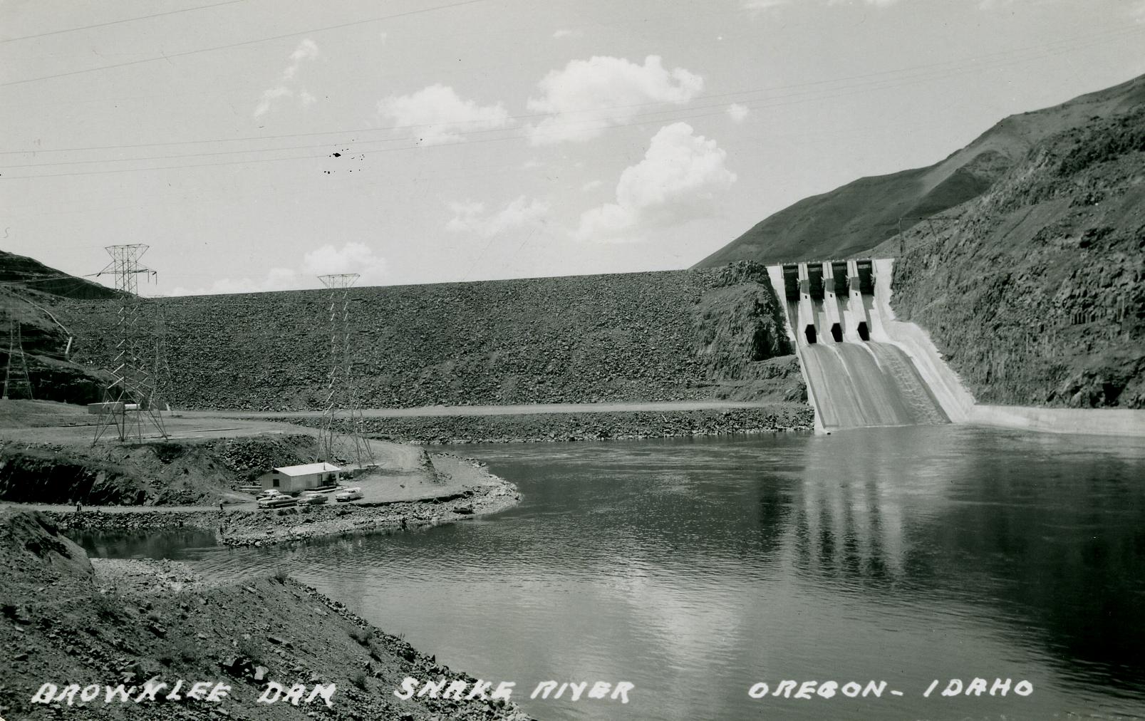 Black-and-white photograph of Brownlee Dam on the Snake River, Idaho-Oregon border. The photo shows a gravel dam view looking upstream. A narrow concrete spillway occurs on the right side of the dam. Some power lines can be seen on the left riverbank.
