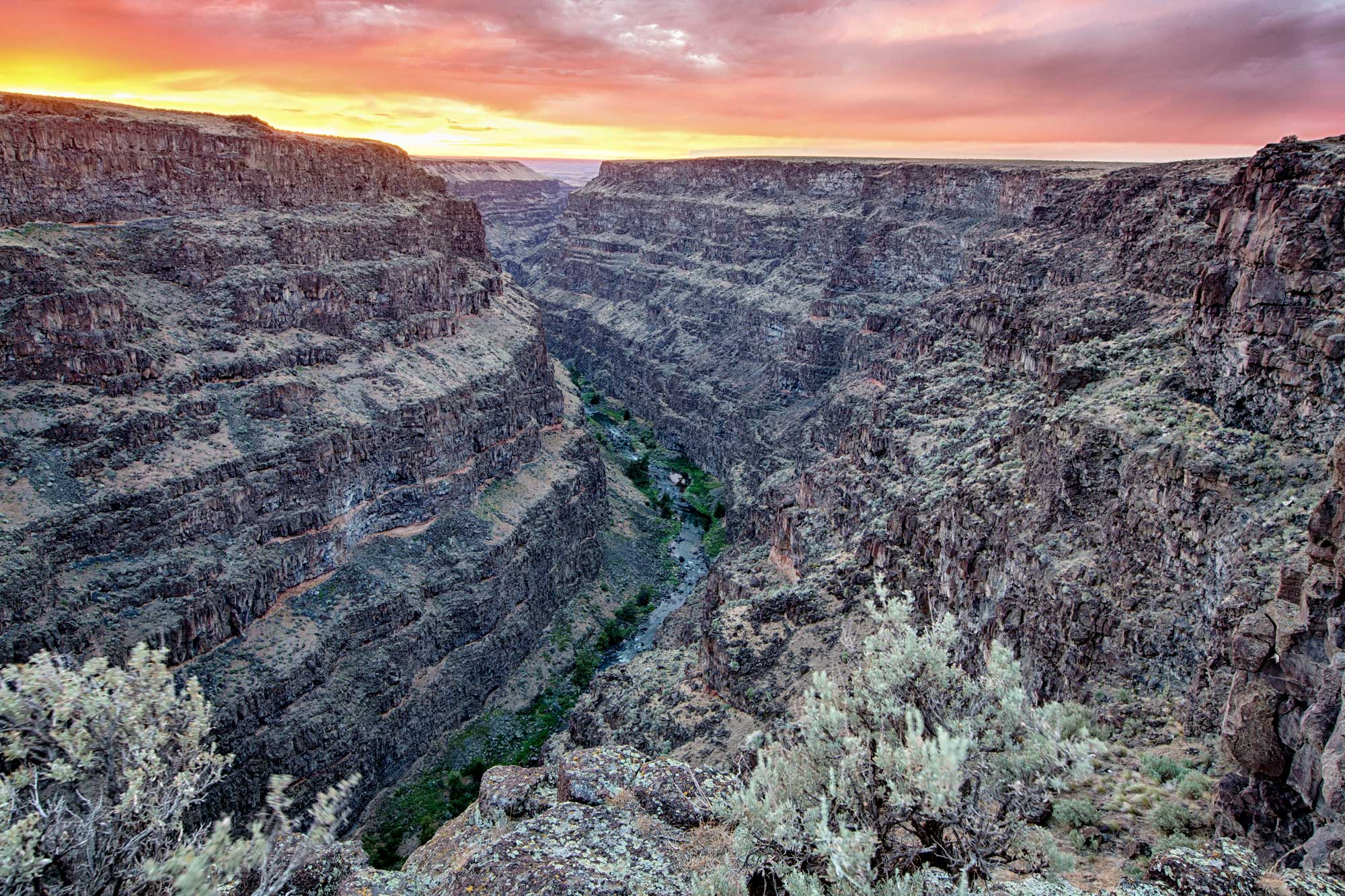 Photograph of a canyon on the Bruneau River in southwestern Idaho. The photo shows a river running through a canyon with steep walls. The walls rise in a steep stair-step fashion on either side of the river. The sky is yellow and orange, perhaps indicating that the photo was taken at sunset.