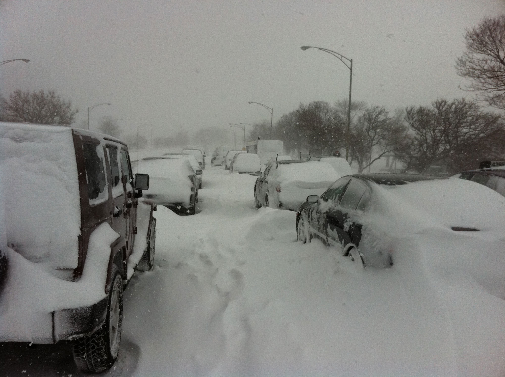 Photograph of Lake Shore Drive in Chicago during a snowstorm in February 2011. The photo shows a road extending from the foreground into the distance. The road has two rows of cars. Both road and cars are covered with snow. Snow filled footprints can be seen between the rows of cars. The sky is grayish-white and snow is falling.