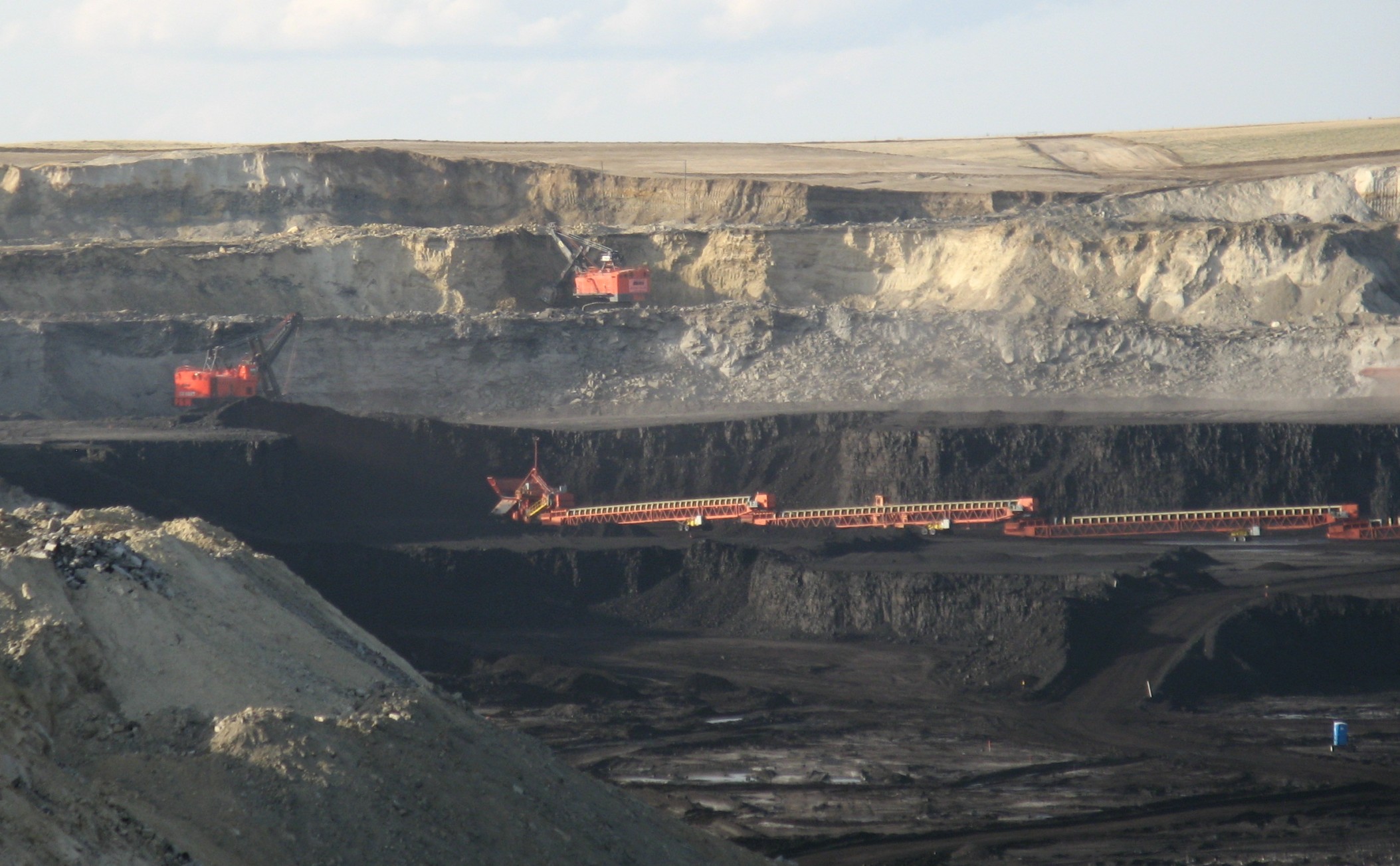 Photograph taken in a coal mine in the Powder River Basin of Wyoming. The photo shows the wall and floor of an open-pit mine with several levels. Mining machinery, including two draglines, can be seen on three of the levels; the machinery is pained orange. The lower levels of the mine appear black, the upper levels are light gray to beige.
