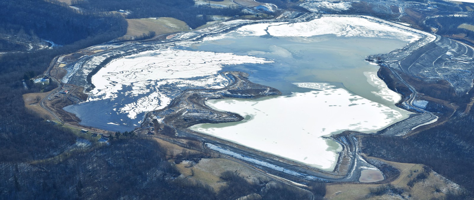 Aerial photograph of a coal slurry pond in Alledonia, Ohio. The photo shows a large, irregularly shaped pond in a forested landscape. Large snow patches cover the surface of the pond in places.