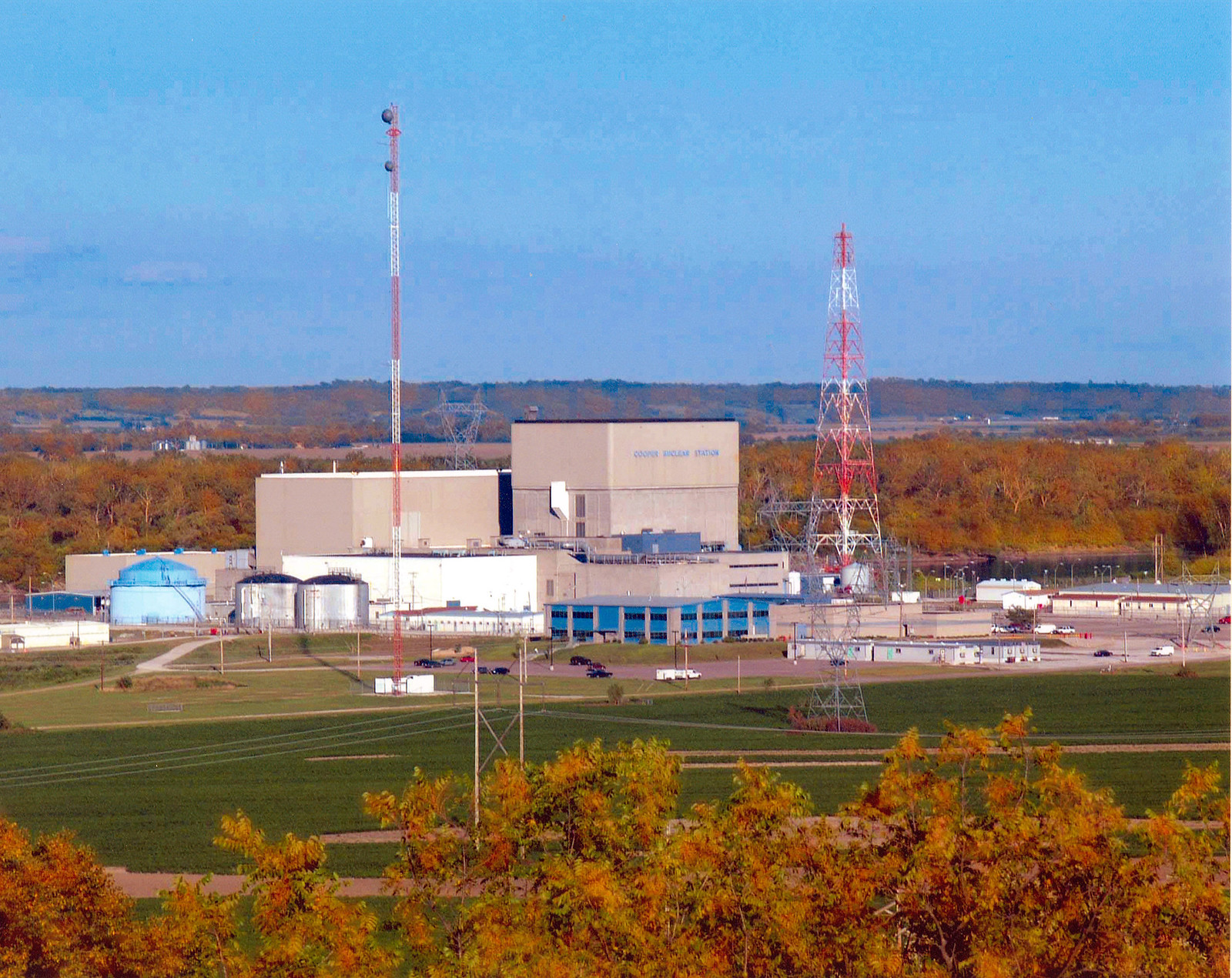 Photograph of Cooper Nuclear Station in Nebraska. The photos shows three squarish brick building of different heights arranged in a stair-step fashion. Around the buildings are tanks, other buildings, and antenna towers. The landscape around is forested, with trees in autumn colors.