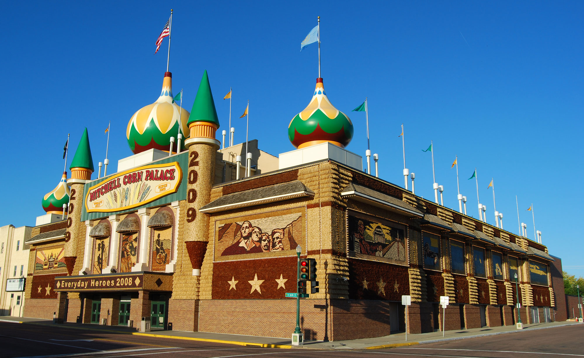 Photograph of the Corn Palace in Mitchell, South Dakota. The photo shows a rectangular building with conical and onion-dome-topped towers. The building is decorated in beige, burgundy, green, and yellow. The two towers with conical peaks flanking the front entrance say "2009." Flags fly from the tops of the three onion-dome-topped towers on the front corners and in the center-front of the building. Murals on the walls of the building depict various scenes, the the nearest mural showing the faces on Mount Rushmore. The theme of the palace is "Everyday Heroes 2008" as indicated on the marquee overhanging the front doors.