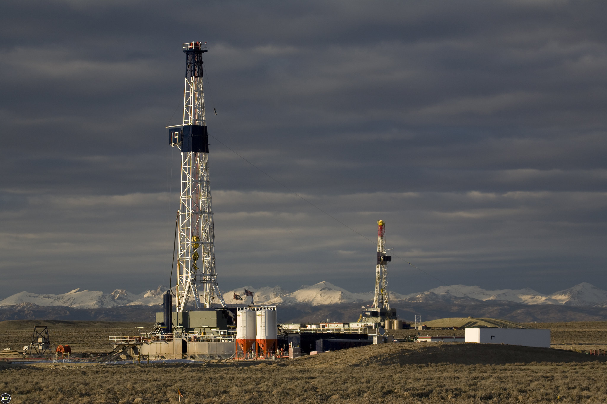 Photograph of drilling rigs in the Pinedale Anticline Natural Gas Field, Wyoming. The photo shows two drill rigs, one in the foreground, one in the background, with associated equipment. The rigs are on a flat, scrubby landscape. Snow-capped mountains can be seen on the horizon. The sky is overcast.