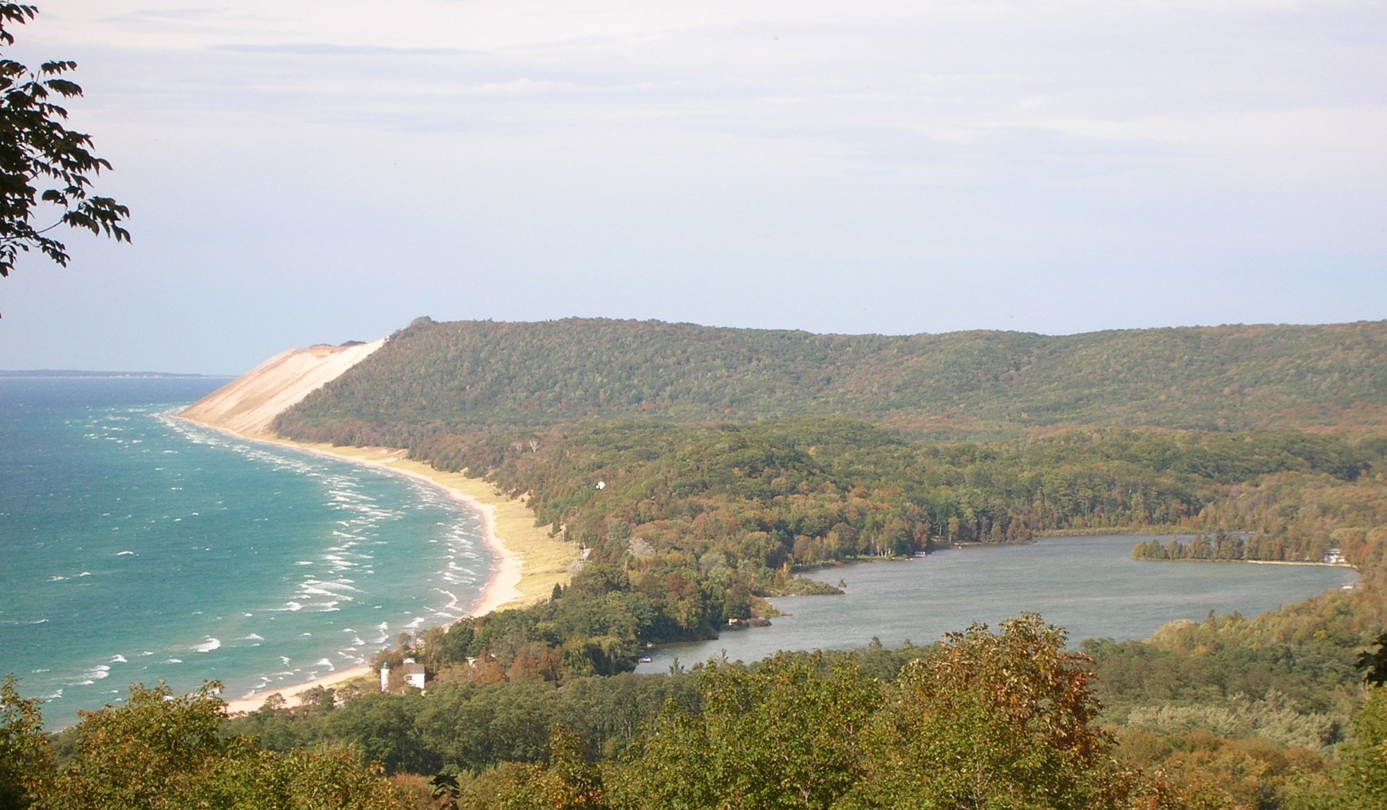 Photograph of Empire Bluff in Sleeping Bear Dunes National Lakeshore. The photo shows the arching shoreline of Lake Michigan at left, with a sandy bluff rising above the lake in the distance. To the right is a forested landscape with a large lake in a low spot.