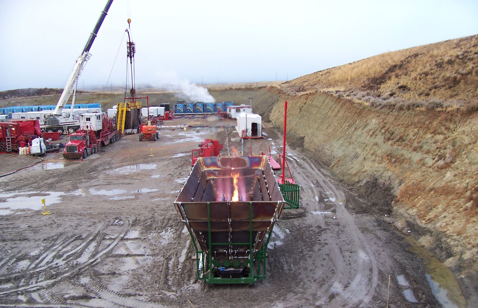 Photograph of an excavated area in the Pinedale Anticline Natural Gas Field. An enclosed gas flare is in the foreground. It looks like a metal container with a flame inside. In the background, vehicles and other equipment are visible.