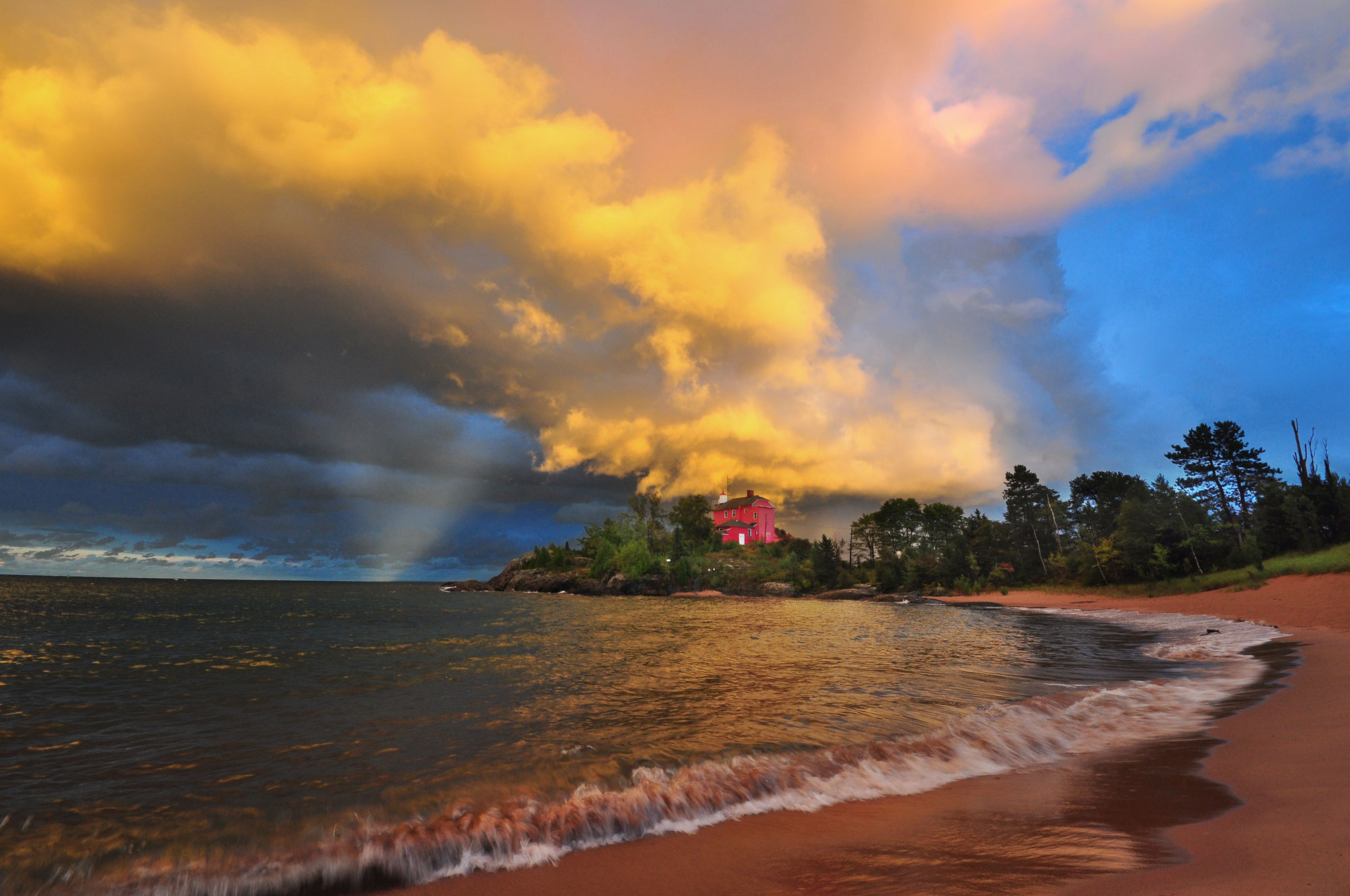 Photograph of the shore of Lake Michigan in Marquette, Michigan during the fall. The photo shows the shoreline of the lake arcing away from the center foreground, with a narrow sandy beach on the right and water on the left. In the distance, a red building stands on a low point of land, where it is surrounded by trees. The sky above is blue with fluffy clouds lit dramatically by the sun.