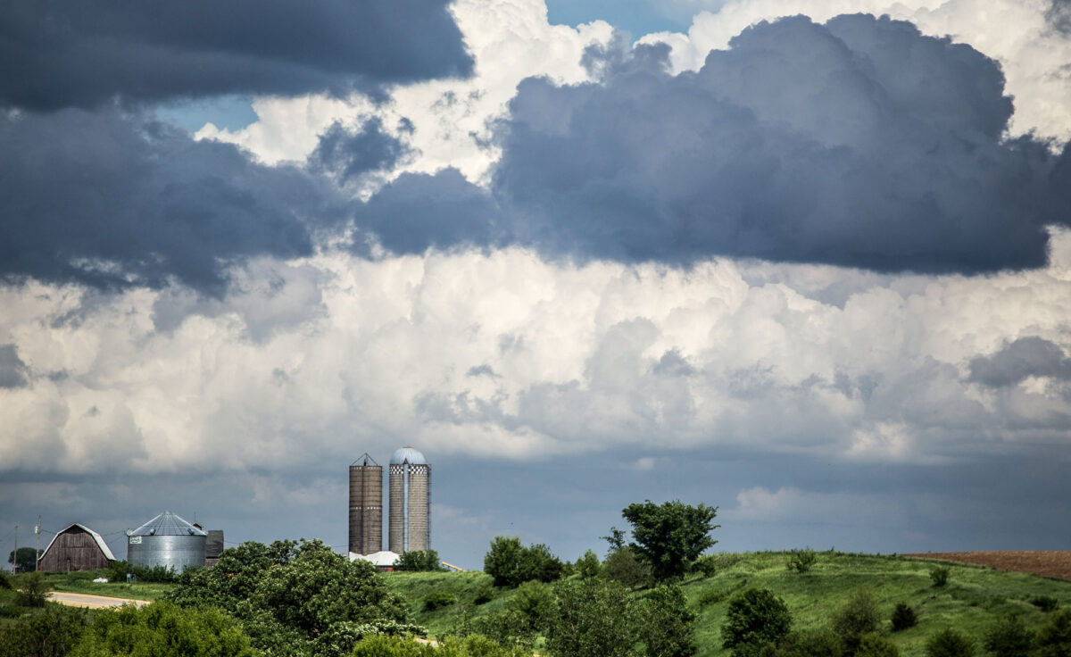 Photograph of a farm in Iowa on a summer day. The photo shows a slightly hilly landscape with green grass and trees. Part of a brown field can be seen to the right. To the left are silos and a barn. The sky above the field dominates the image. It is overcast with fluffy gray and white clouds.
