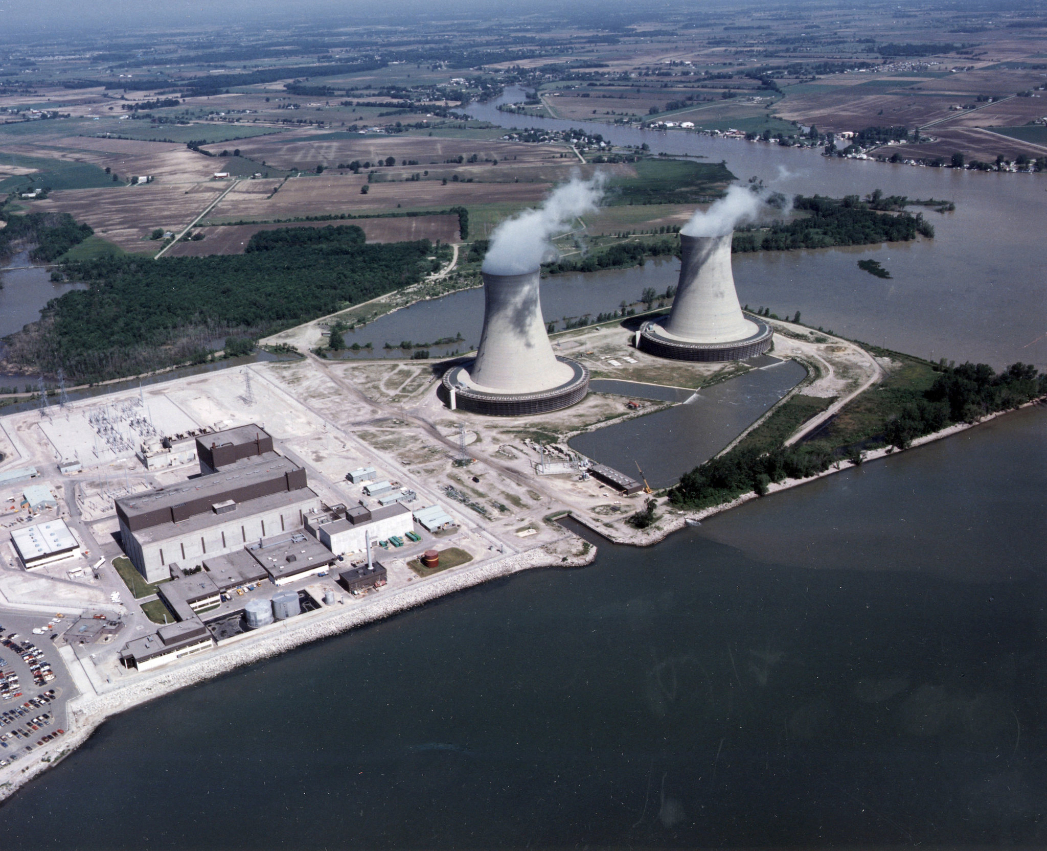 Aerial photograph of Enrico Fermi Nuclear Generating Station in Newport, Michigan. The photo shows two large cooling towers on a point extending into water. Nearby is a rectangular bulidng and other structures. In the distance, the flat landscape is made up of a patchwork of fields and trees.