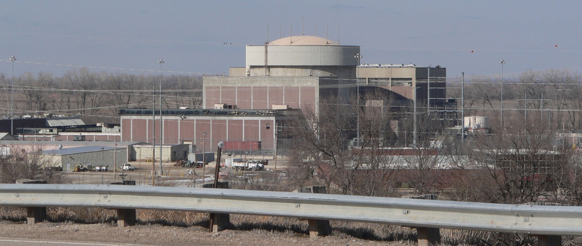 Photograph of Fort Calhoun Nuclear Generating Station in Nebraska. The image shows a large brick and concrete building with several levels, the highest appearing to be a cylindrical structure capped by a convex roof. The landscape around the station is forested, the leafless trees (indicating that it is probably winter).