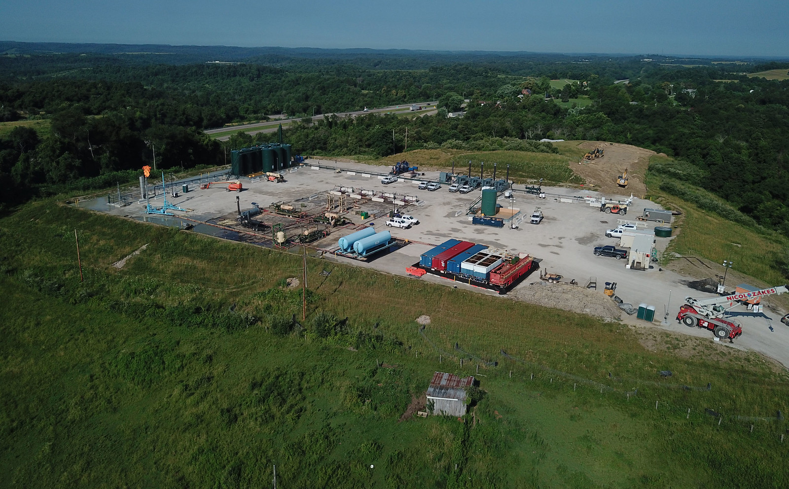 Aerial photograph of a fracking pad, Fairview, Ohio. The photo shows a flat, cleared space on top of a hill. The cleared space has equipment and machinery on it. The surrounding landscape is green and covered with trees, grass, and other plants.