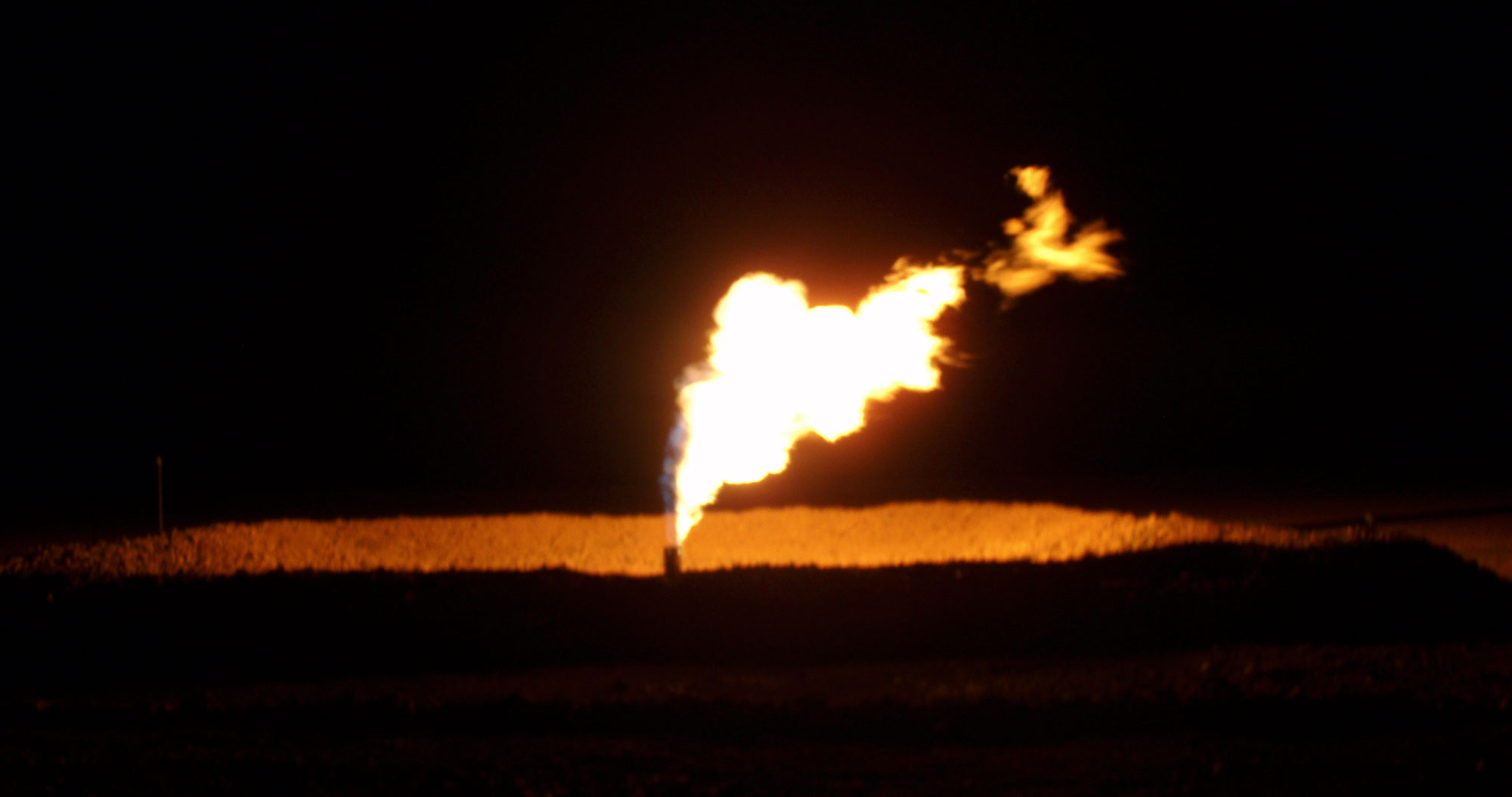Photograph of a gas flare in the Bakken Formation of North Dakota at night. The photo shows flame come from the end of a pipe. The remainder of the landscape is mostly dark.