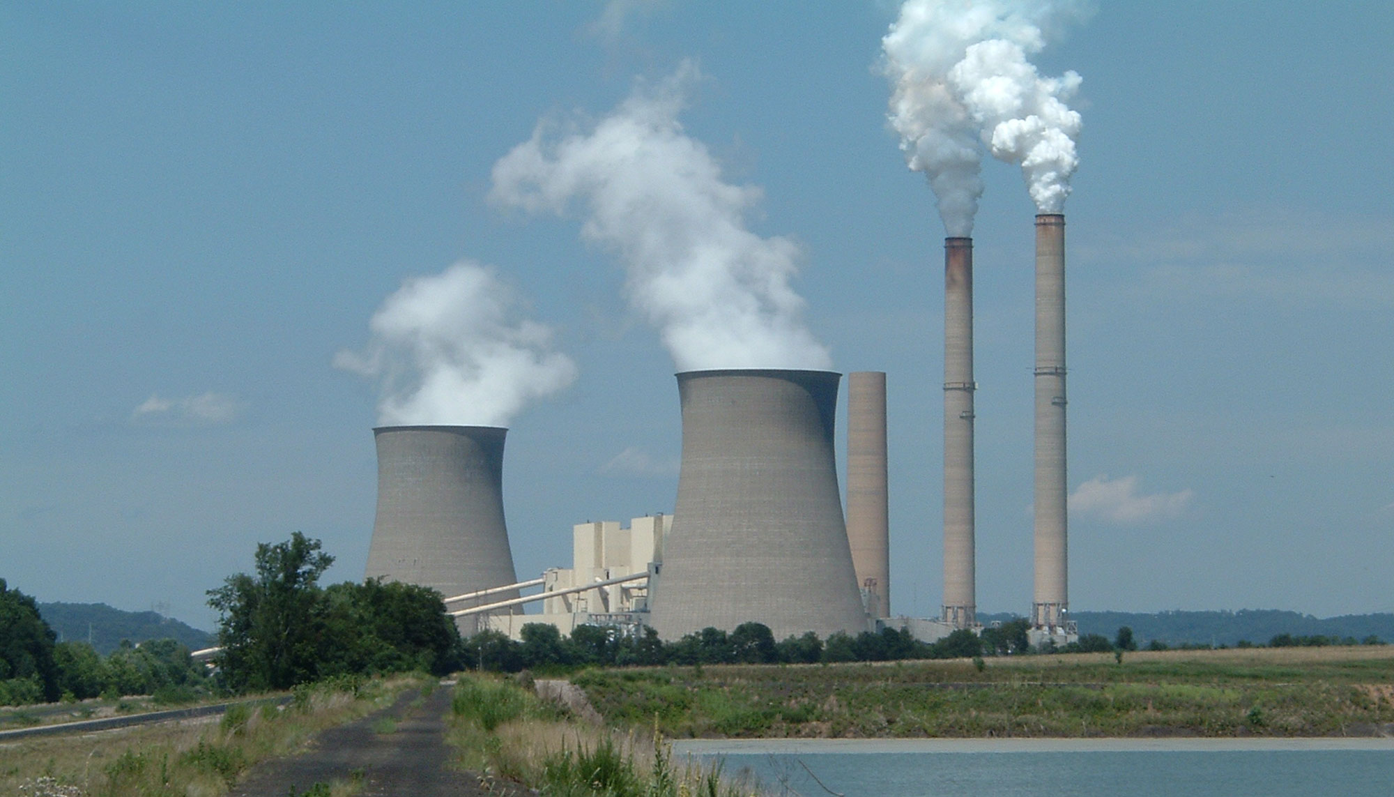 General James M. Gavin Plower Plant in Cheshire, Ohio. The photo shows two large cooling towers and three smokestacks sitting in a flat field. A beige building appears to be situated between the cooling towers. In the foreground, a two-track road runs toward the power plant. To the right of the road is water.