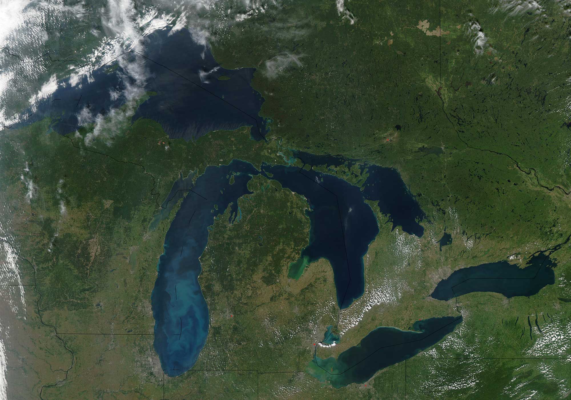 Satellite photograph of the Great Lakes in the midwestern and northeastern United States. The photo shows the five Great Lakes, from west to east, Superior, Michigan, Huron, Erie, and Ontario. The lakes are dark blue and surrounded by green landscape. The green generally becomes darker to the north, indicating more forest cover.