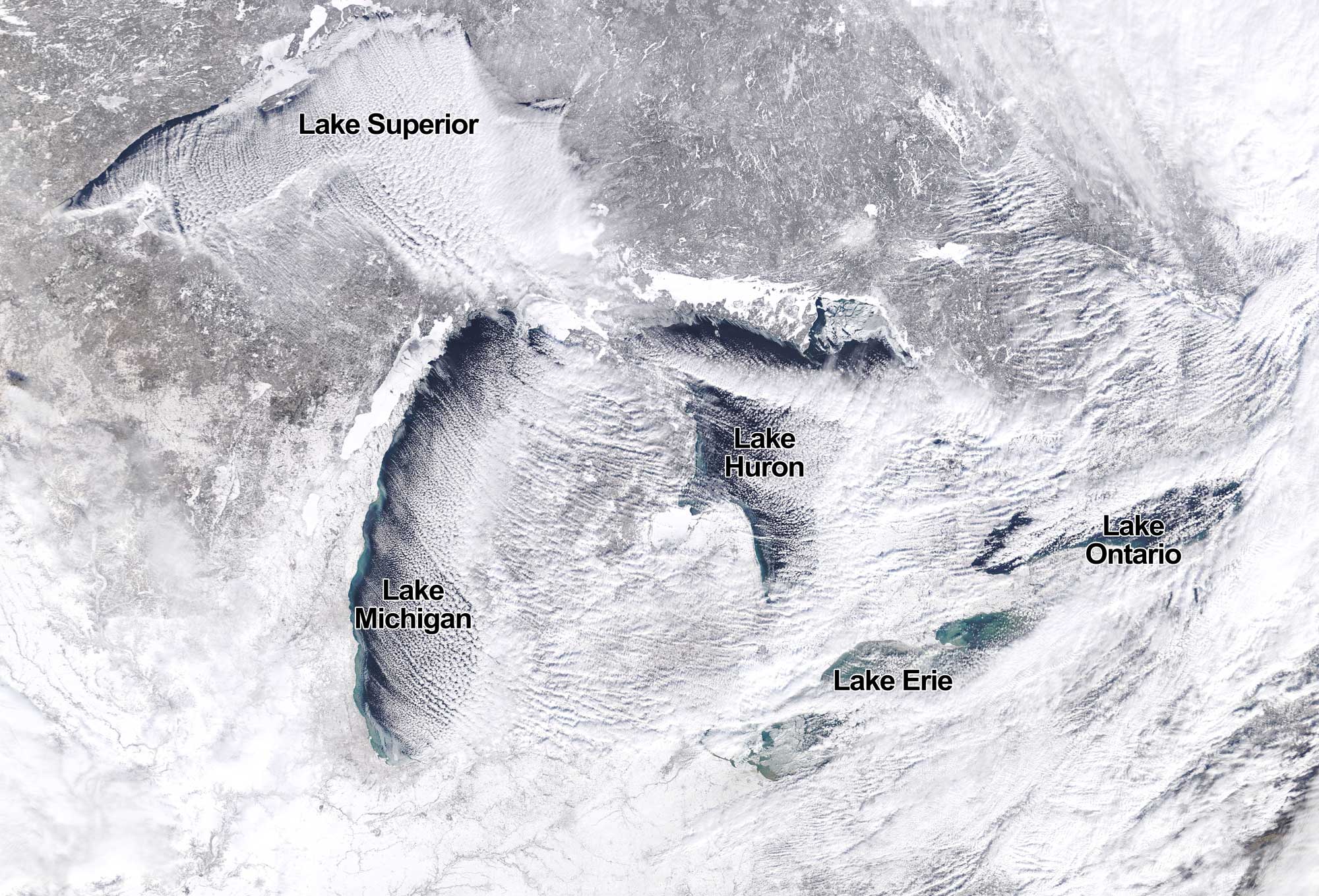 Satellite photo fo the Great Lakes region during the winter with all five lakes labeled. The photo shows that the land is dusted by white snow. Clouds stream from above Lake Superior and Lake Michigan in curved parallel rows traveling to the southeast. These clouds carry lake-effect snow.