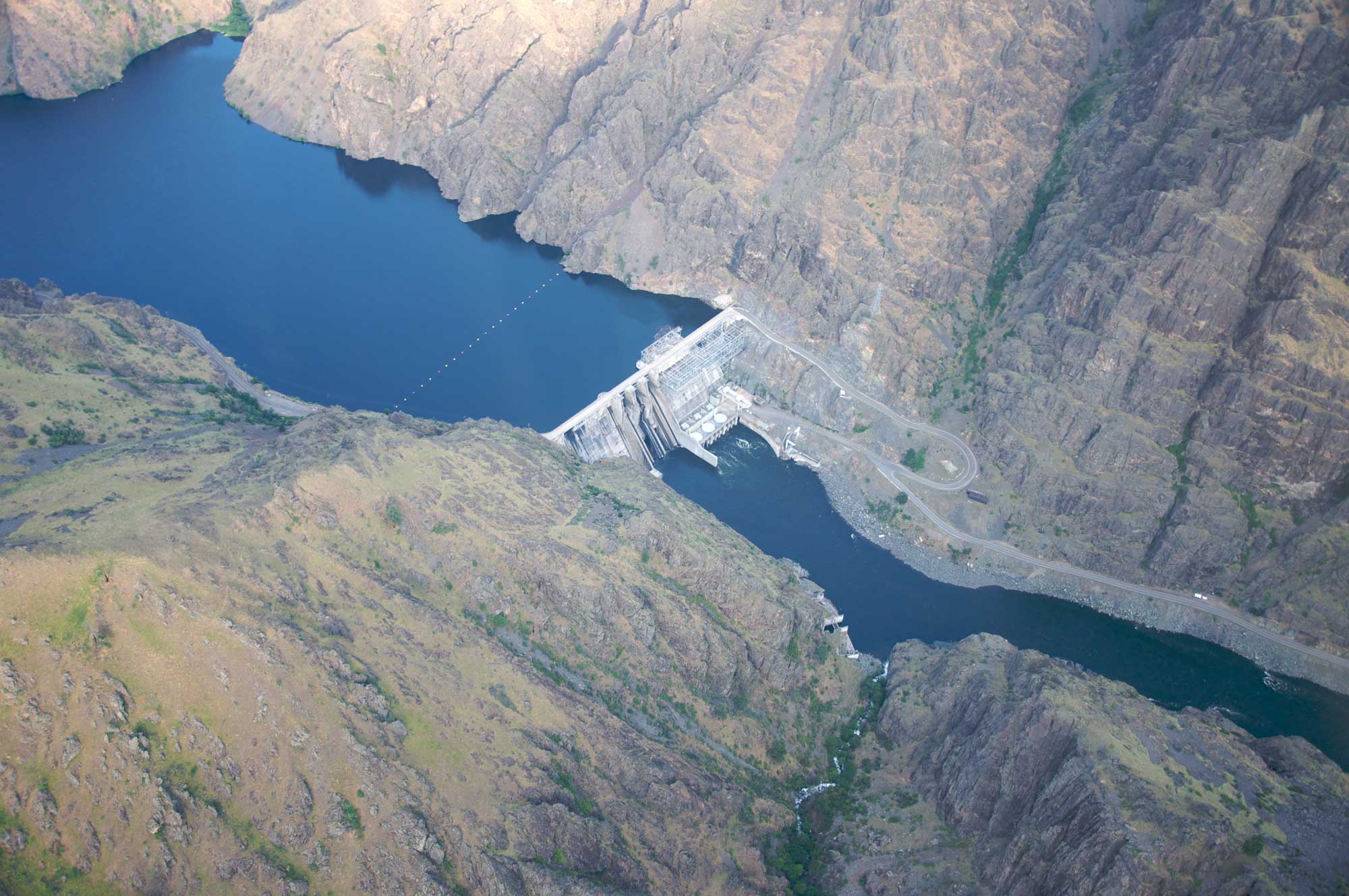 Aerial photograph of Hells Canyon Dam on the Snake River on the Idaho-Oregon border. The photo shows a concrete dam built across a river at the bottom of a canyon with steeply sloping walls. A reservoir is held back behind the dam, whereas a river can be seen downstream from the dam. On the far wall of the canyon, a road can be seen leading up to the dam.