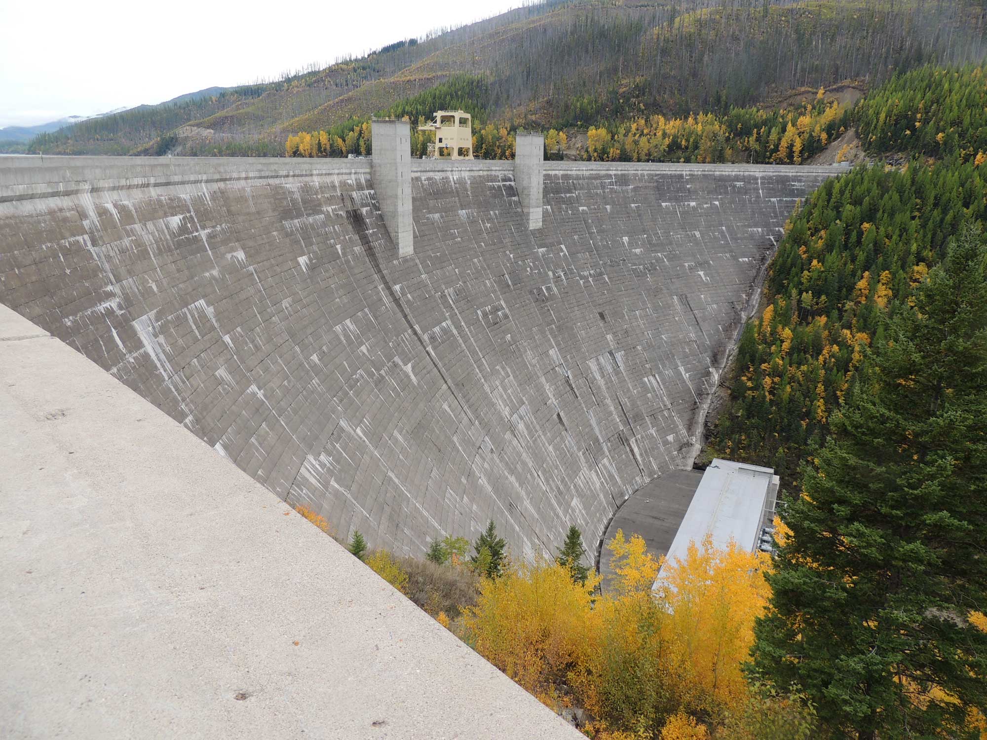 Photograph of Hungry Horse Dam on the Flathead River in Montana. The photo shows a tall concrete dam. The photo is taken from one side of the dam looking across to the opposite shore. Conifer-covered slopes rise in the distance, presumably next to a reservoir.