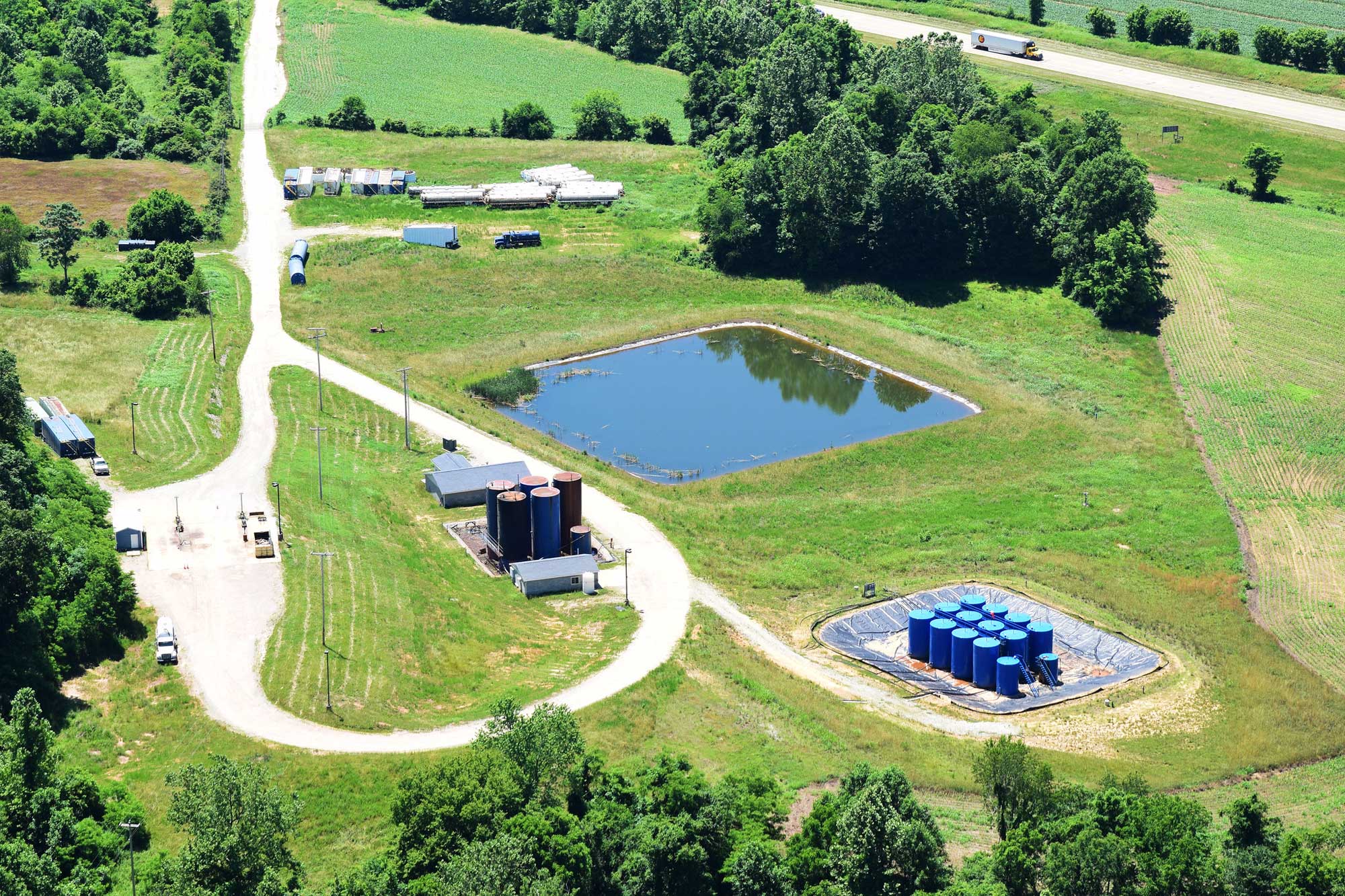 Aerial photograph of an injection well site in Meigs County, Ohio. The photo shows a road ending in a loop, with a series of tall storage tanks on the inner edge of the right side of the loop. To the right, a spur road leads to another group of storage tanks. A square holding pond is in the center of the image. The surrounding landscape green with grass, trees, and scattered groups of structures.