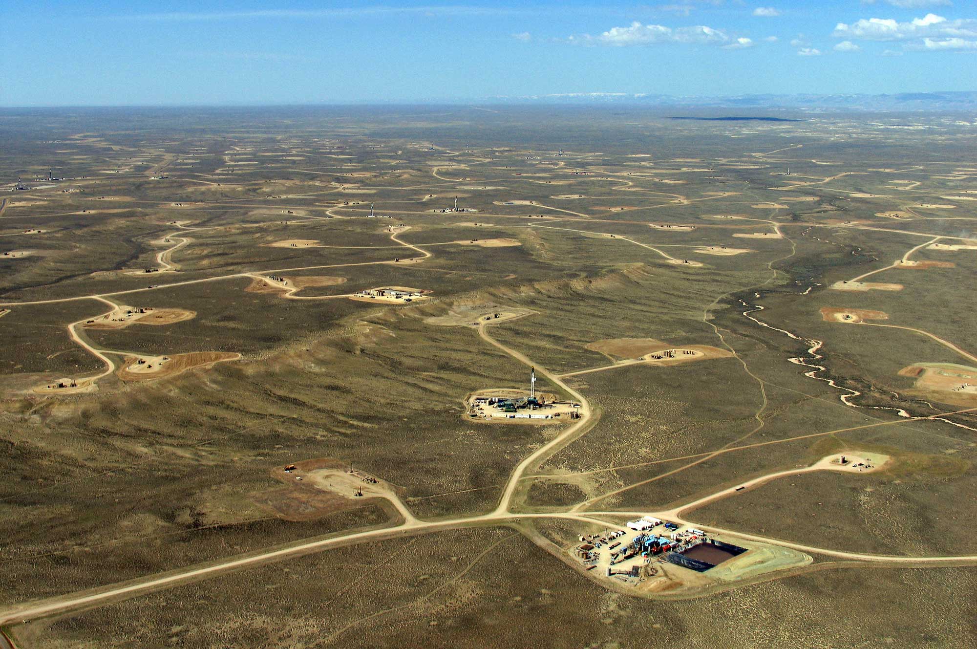 Aerial photograph of the Jonah Gas Field in Wyoming. The photo shows a flat, brown landscape with a network of dirt roads that have branches that end in oil well pads, cleared rectangular areas that support drilling and pumping equipment.