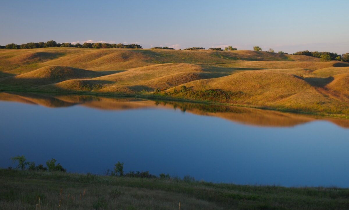 Photograph of Kettle Lake in Glacial Lakes State Park, Minnesota. The photo shows part of a lake with still water in the foreground with gently rolling hills in the background. The hills are covered with grass. Trees can be seen on the horizon.