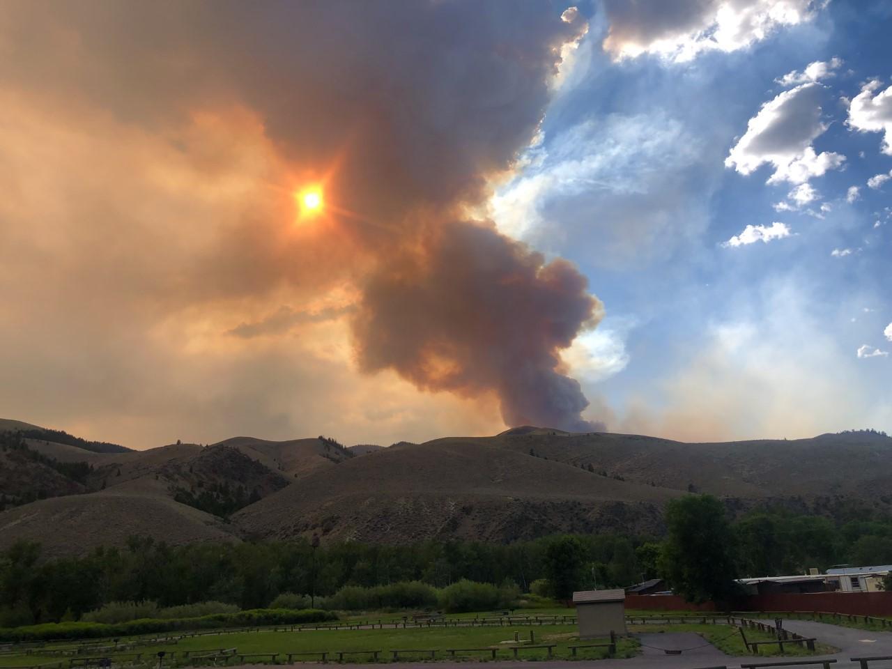 Photograph of the Moose Fire in Idaho, 2022. The photo shows a plume of smoke rising from behind rolling hills. The sun can be seen through the smoke, and it appears orange. The part of the sky not blotted out by smoke in the upper right side of the image is blue with fluffy clouds. The foreground is a flat landscape with green vegetation.