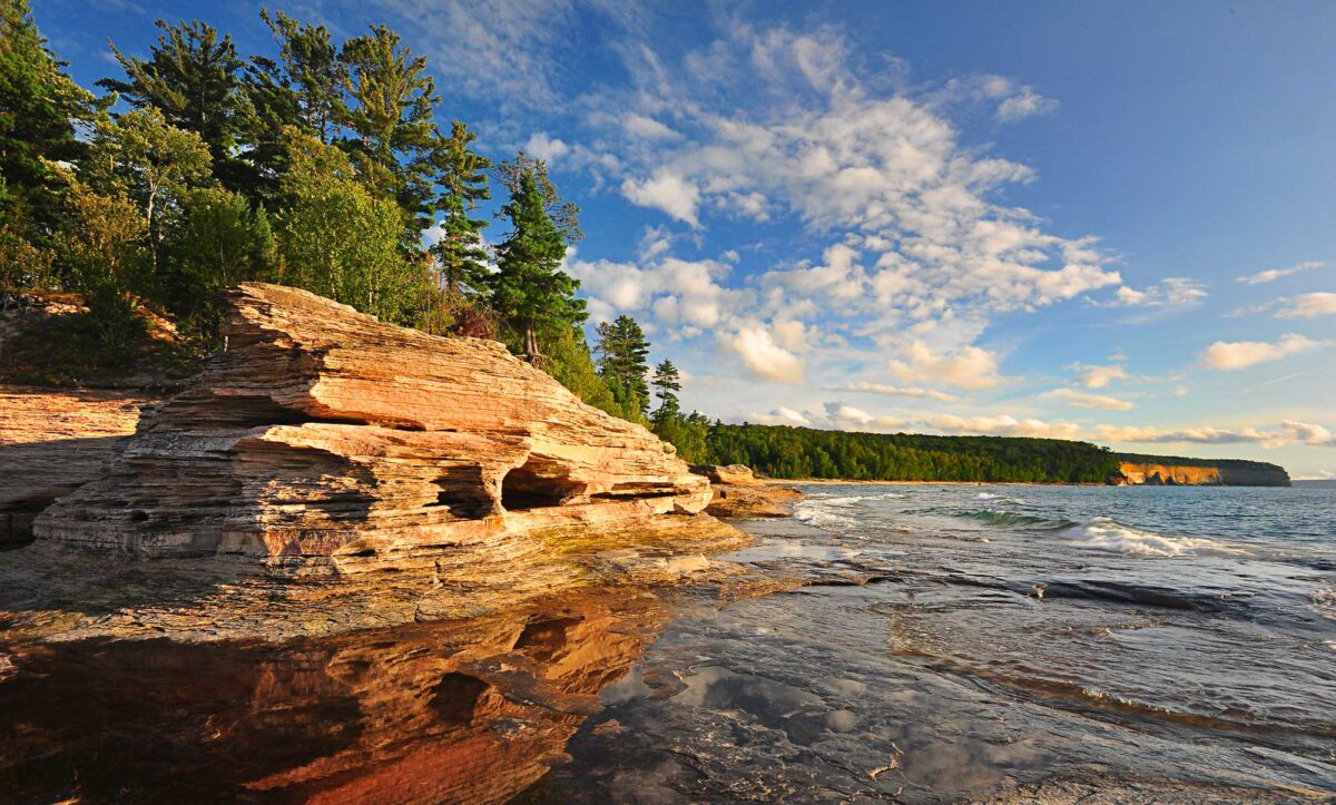 Photograph of Mosquito Beach at Pictured Rock National Lakeshore, Lake Superior, Michigan. The photo shows a weathered rock formation made out of orangish-beige stone at the left foreground. Trees are growing on top of it, and it is reflected in the water at its base. In the background, more of the lakeshore can be seen stretching into the distance.