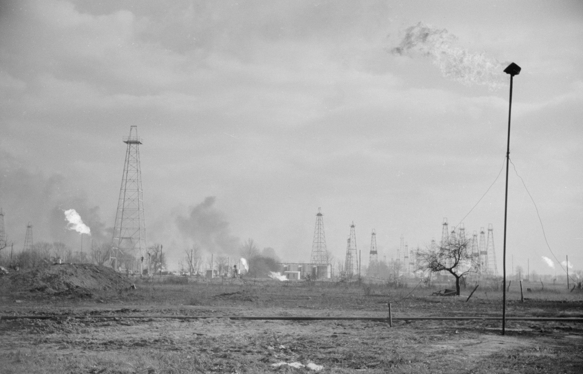 Black and white photo of an oilfield in Marion County, Illinois, in 1940. The photo shows a flat landscape beneath an overcast sky. Oil derricks and gas flares dot the landscapes. Other structures ca be seen in the far distance behind some derricks.
