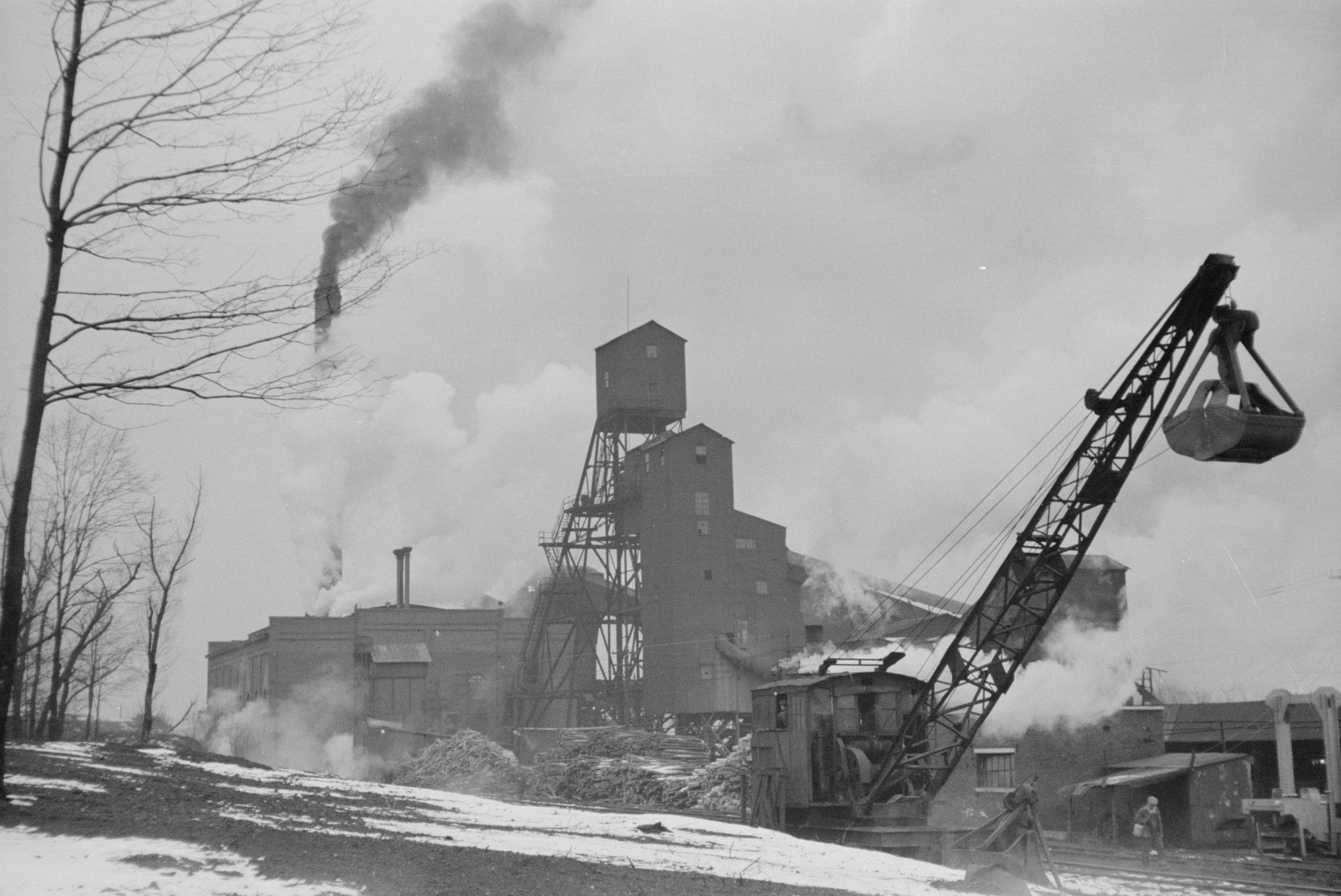 Black and white photo of Orient Mine No. 1 in Franklin County, Illinois, in 1939. The photo shows a a tall, possibly wooden structure in the background, in front of another building from which smokestacks are belting dark gray and white smoke. There is a large pile of logs in front of the building, and in the foreground in a large dragline excavator. Other building can be seen at right, with two people walking in front of them. The trees at right are barren of leaves and the ground has snow on it, indicating that it is winter.