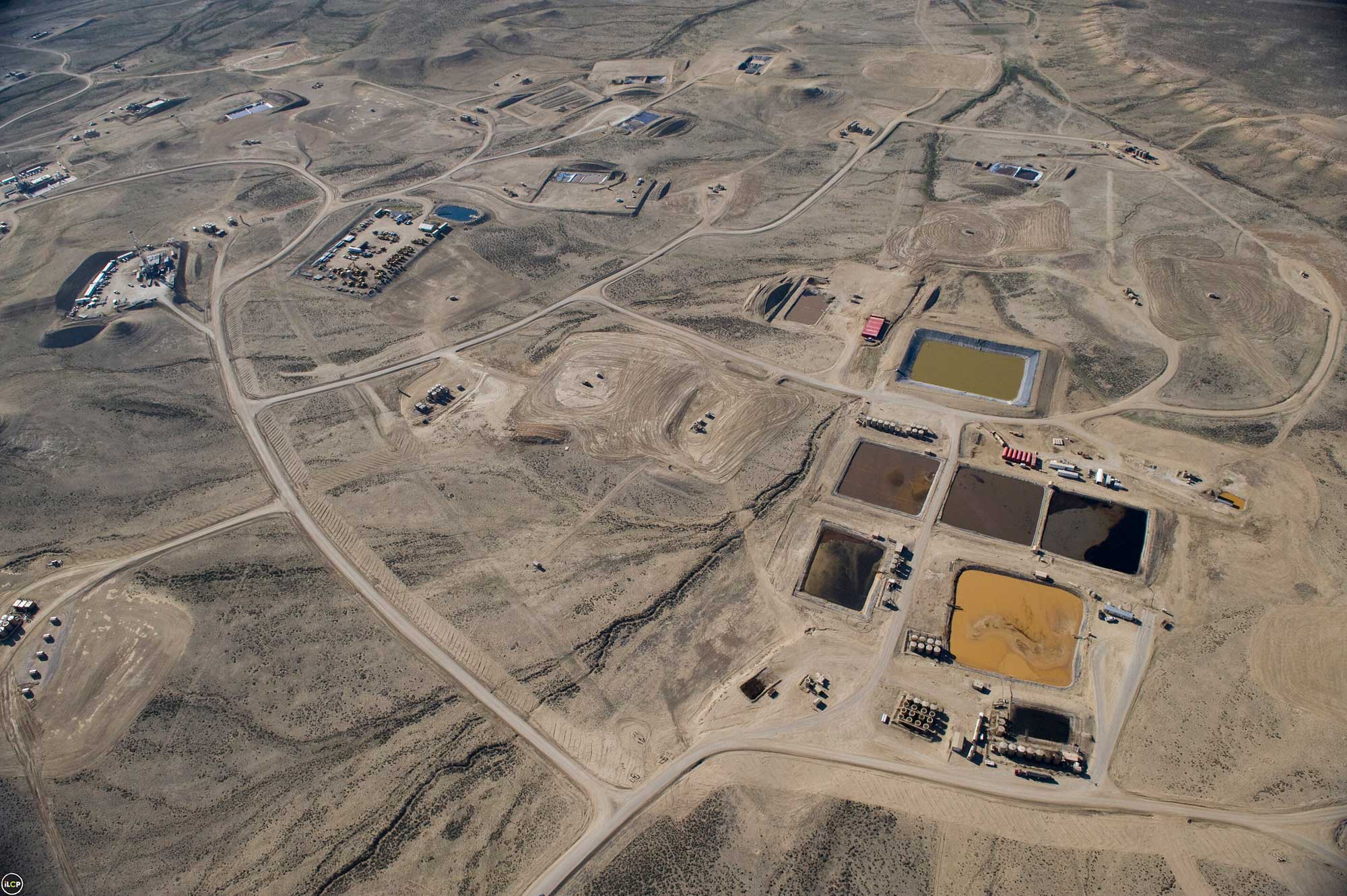 Aerial photograph of the Pinedale Anticline Natural Gas Field, Wyoming. The photo shows a brown landscape with a network of roads and oil well drilling pads. In the foreground at right, a series of square to rectangular waste pits can be seen. The pits are open to the air and hold green, orange, and brown liquids.