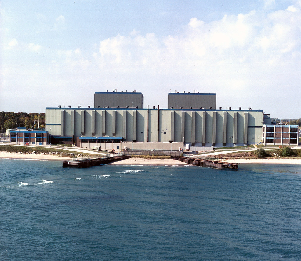 Photograph of Point Beach Nuclear Power Plant in Manitowoc, Wisconsin. The photo shows a long rectangular building with two towers behind it. In front of the building is a narrow sandy beach and the blue water of a lake. 