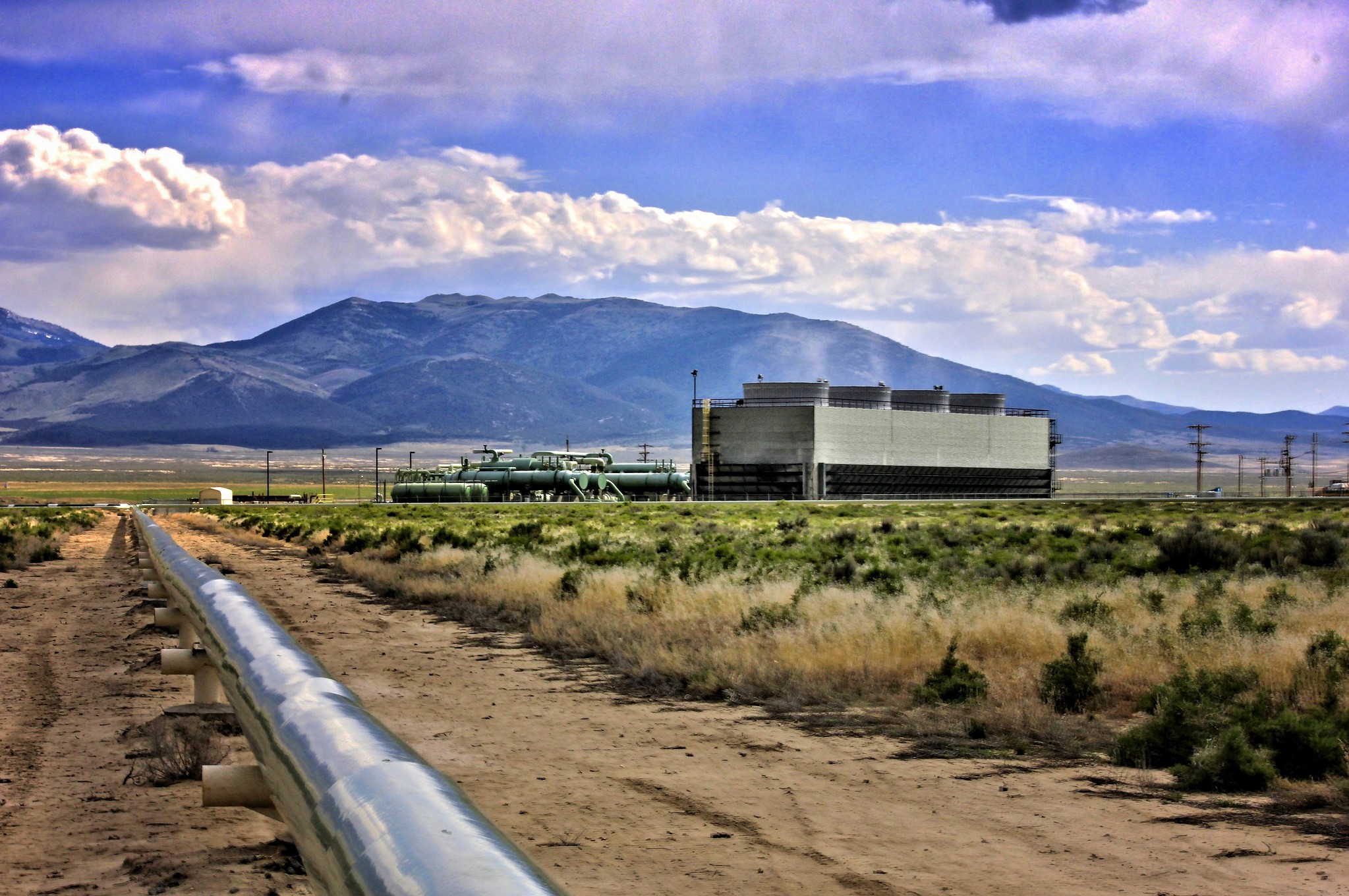 Photograph of the Raft River Geothermal Power Plant in Idaho. The photo shows a rectangular structure with four short cylindrical structures on top of it. Smoke appears to be coming from the cylinders. Green tanks can be seen next to the rectangular structure. A pipe runs from the geothermal plant to the foreground of the image. The landscape is flat with scrub. Low mountains rise in the background with a cloudy blue sky above.