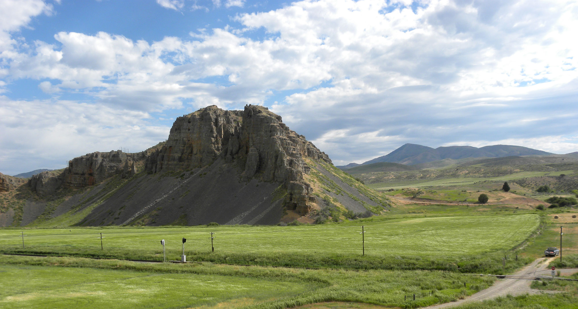 Photograph of Red Rock Pass, Idaho. The photo shows a rock formation rising in the middle of a flat green field. More low hills rise in the background. A gravel road can be seen in the lower right corner of the image.