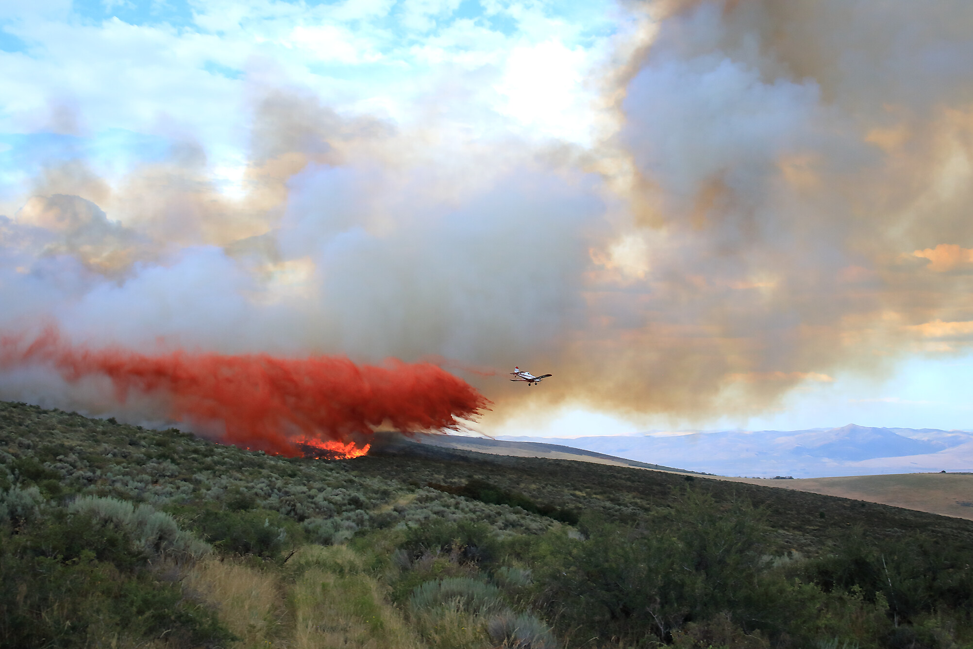 Photograph of an airplane dropping fire retardant on the Ross Fork Fire in Idaho, 2022. The photo shows a small white plane flying over a hilly landscape covered with scrub. On the nearest hill, orange flames from a fire can be seen. The plane is dropping a red substance over the flames. Gray-brown smoke can be seen in the background.