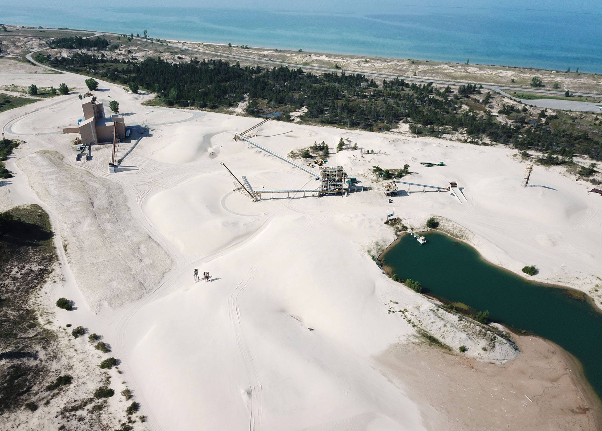 Photograph of a sand dune mine in Michigan. The photograph shows mining structures on a dune made up of off-white sand. In the lower right corner of the image is a pond. In the background, from upper left to slightly lower on the right is the Lake Michigan shoreline. The sand mine is separated from the shore by a patch of trees.