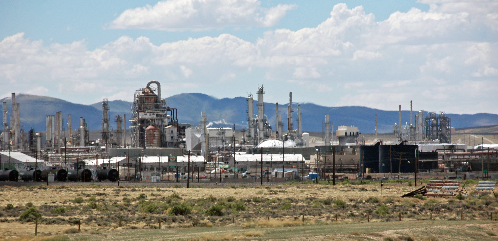 Photograph of the Sinclair oil refinery, Sinclair, Wyoming. The photo shows a cluster of structures in a flat field with hills rising in the background. The sky is blue with scattered clouds.