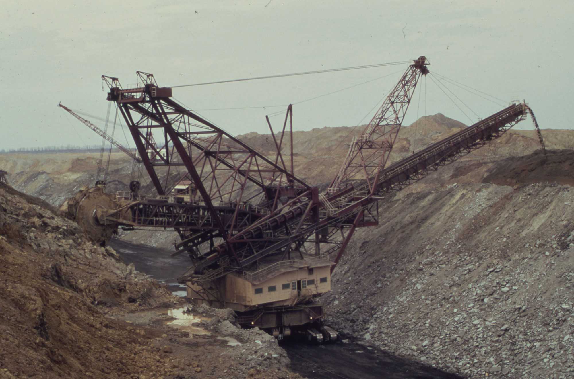 Photograph of a strip mine in Fulton County, Illinois, 1972. The photo shows a bucket wheel excavator in a trench. On the back of the excavator, a wheel with bucket spokes digs into a bank. On the front of the excavator, a conveyer empties sediment at the other edge of the trench. 