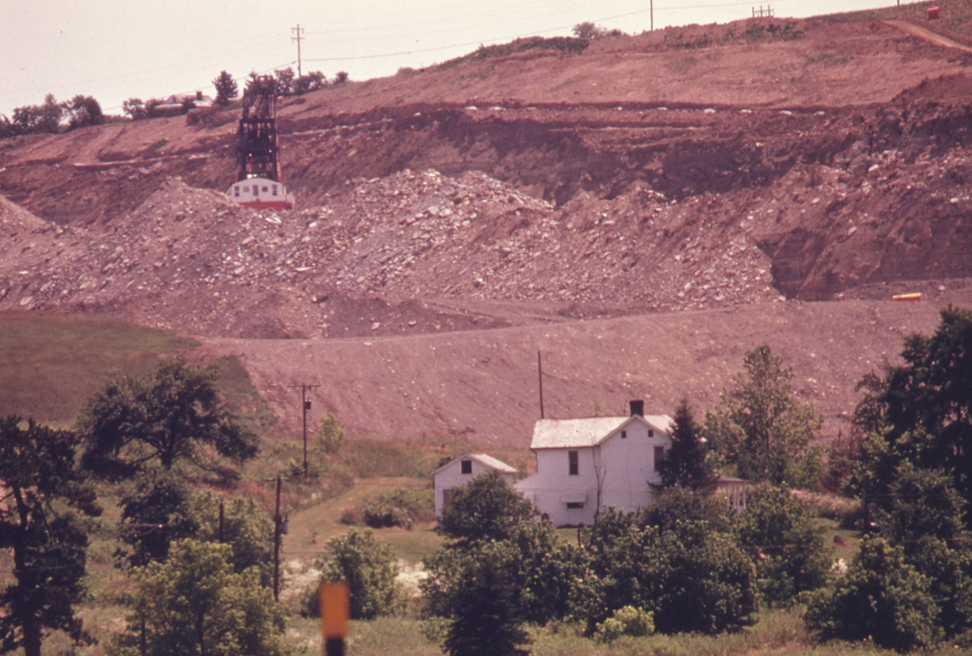 Photograph of a strip mining operation in Ohio in 1974. The photo shows a white house amongst trees in the foreground. A hill rises behind the house in the background. The hill is being excavated by a dragline, which is behind and above the house at the upper left of the image. Large piles of earth can be seen between the house and the dragline.