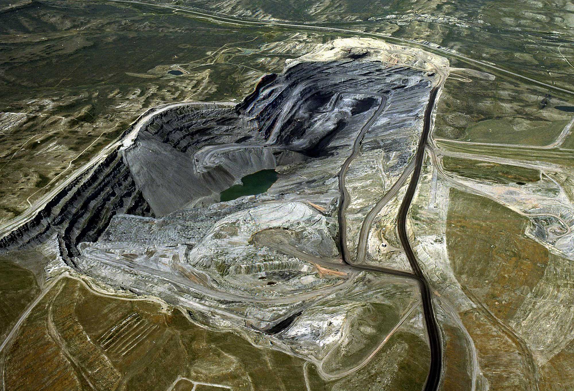 Aerial photograph of an open-pit coal mine in the Powder River Basin of Wyoming. The photo shows a large pit with roads along its rim and also running to it. One side of the pit has terraced (stair-step like) walls. Water has pooled in the deepest part of the pit. 