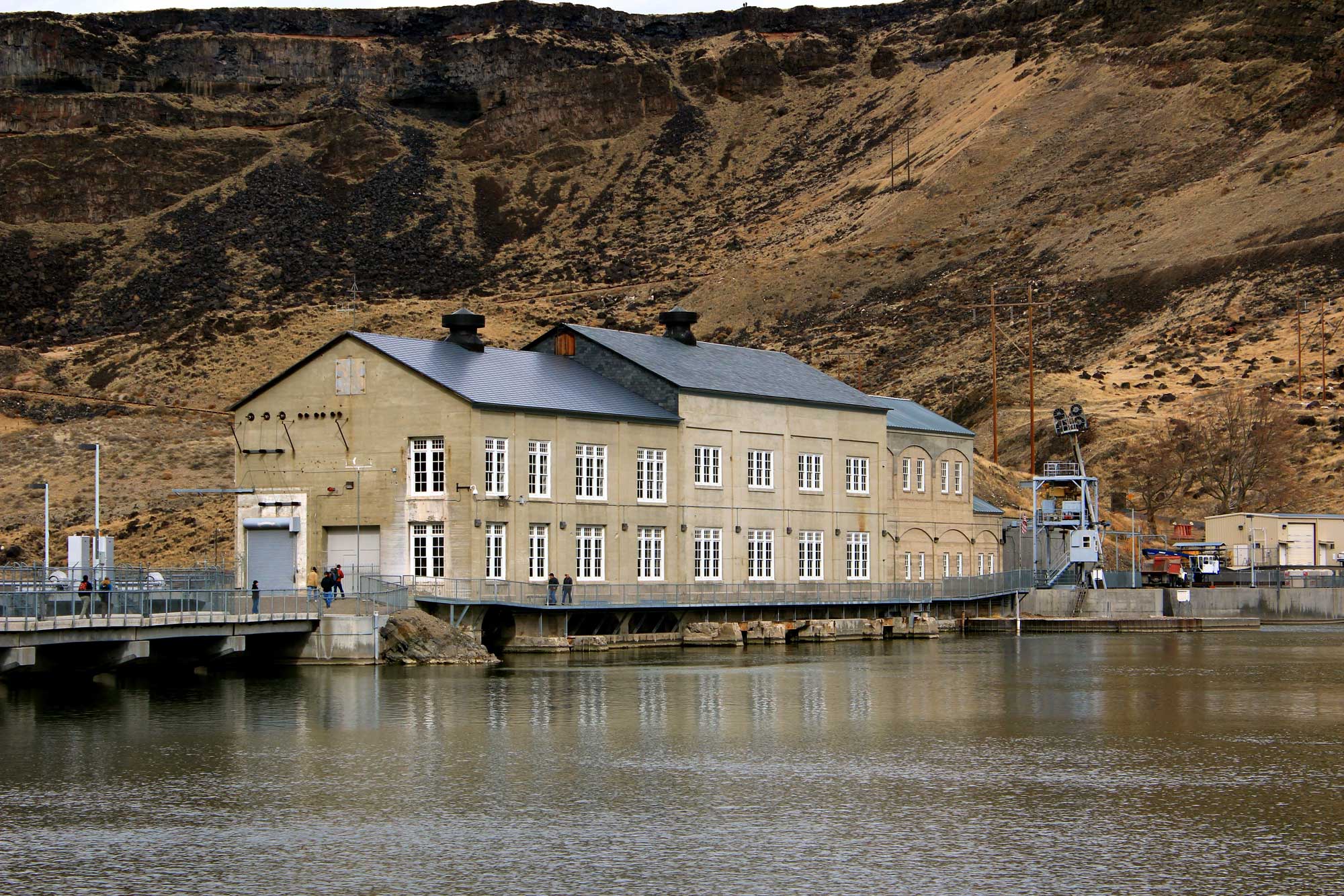 Photograph of Swan Falls dam on the Snake River in Idaho. The photo shows the dam as seen from the shore of the reservoir. A long building with beige walls and a black roof appears to be positioned on top fo the dam. A rocky slope rises in the background.