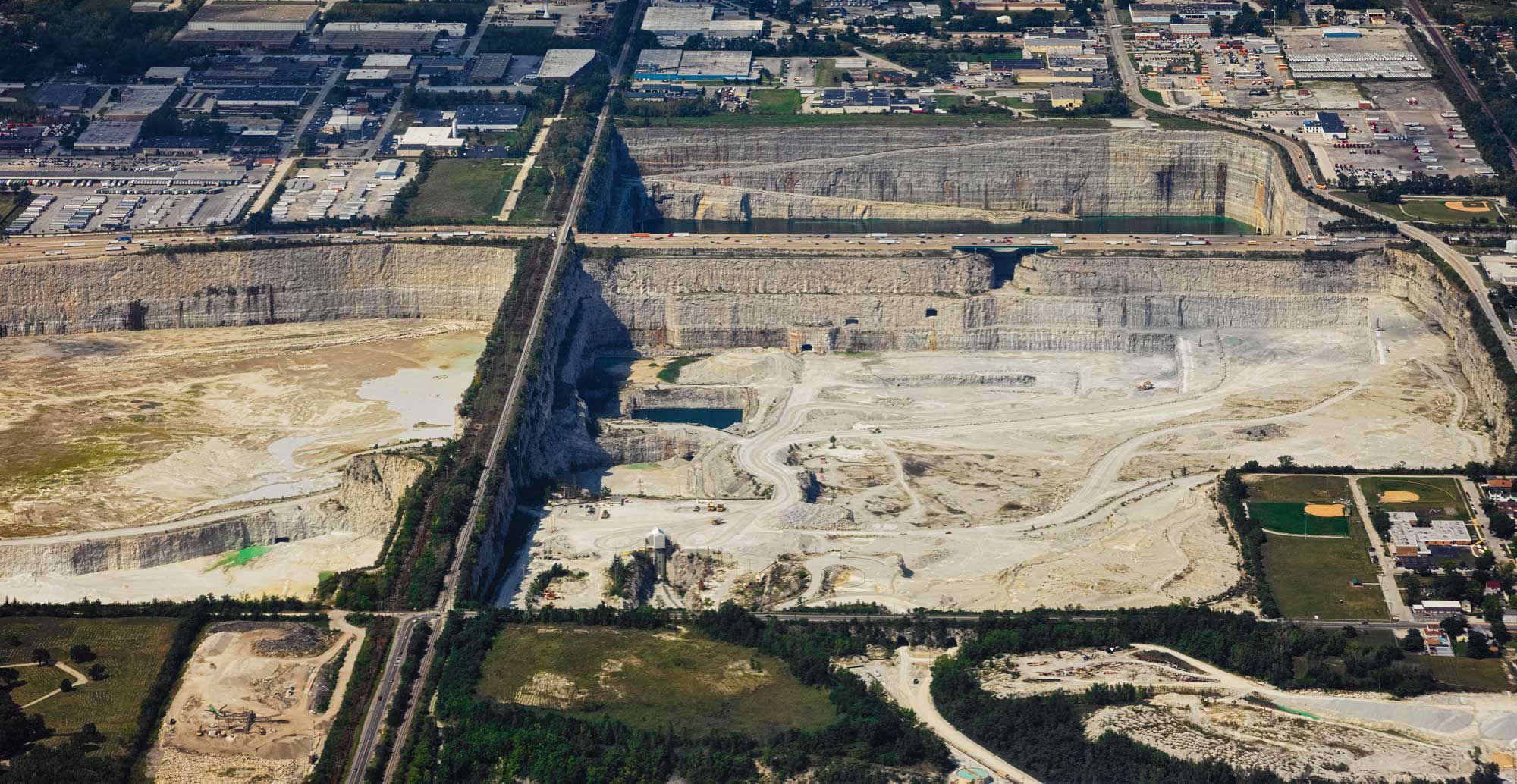 Aerial photograph of Thornton Quarry in northern Illinois. The photograph shows three pits of a large quarry on a flat landscape that is a patchwork of fields and developed areas with buildings and parking lots. The partitions between the quarry pits have roads running on them. The quarries themselves have vertical sides made up of beige rock. Roads and equipment can be seen on the floor of the most visible pit at right foreground. This quarry produces aggregates.