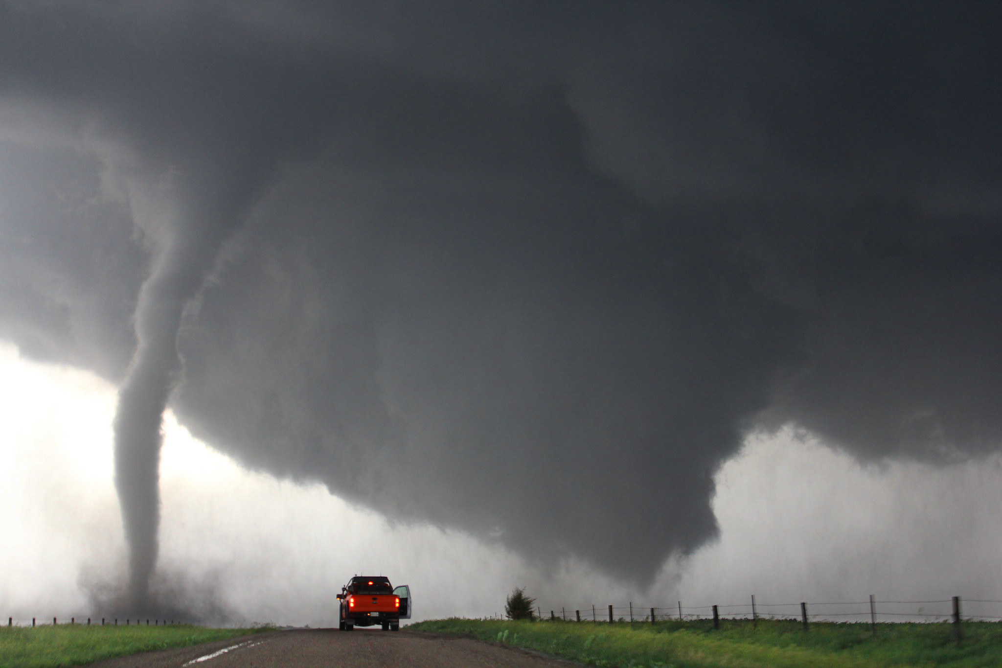 Photograph of a tornado in Nebraska, 2014. The photograph shows a dirt road running toward the horizon. The road is in a field of green grass and is flanked by parallel barbwire fences on either side. A red truck is parked on the road with its taillights illuminated and passenger door open. The sky is gray with dark clouds. To the left, a thin tornado has touched down. To the right, what looks like a much wider tornado is either developing or dissipating. 