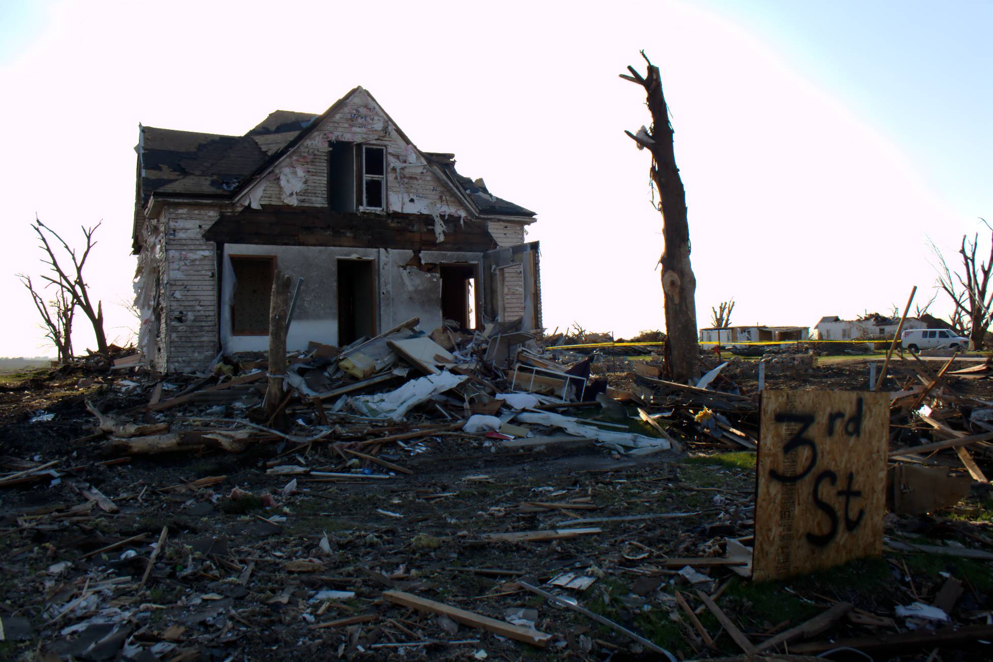 Photograph of a house in Iowa that was damaged by a tornado. The photo shows a house with no windows and doors and damaged siding. The landscape around the house is covered in debris. A particleboard sign in front of the house has "3rd St" spray-painted on it. A tree in front of the house has been stripped of all its branches.